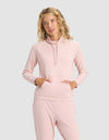 A woman with wavy blonde hair wears the Cozy Earth Women's Brushed Bamboo Mock Neck in light pink, paired with matching pants, standing with her hands in the hoodie pocket against a plain light gray background.|Color:Dusty Rose