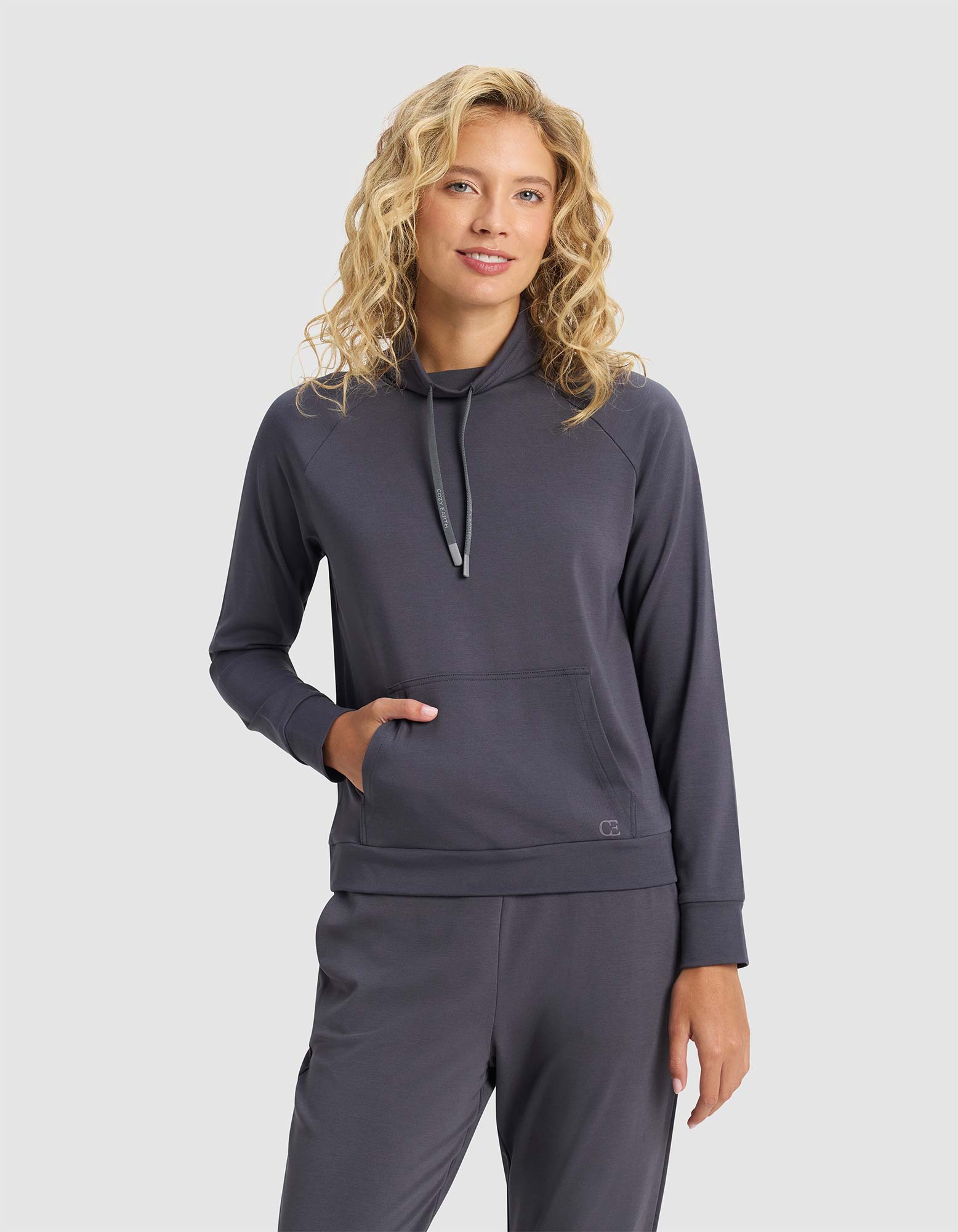 A woman with blonde curly hair wears the Cozy Earth Women's Brushed Bamboo Mock Neck in dark gray, standing against a plain light background with one hand in her pocket and a soft smile.|Color:Graphite