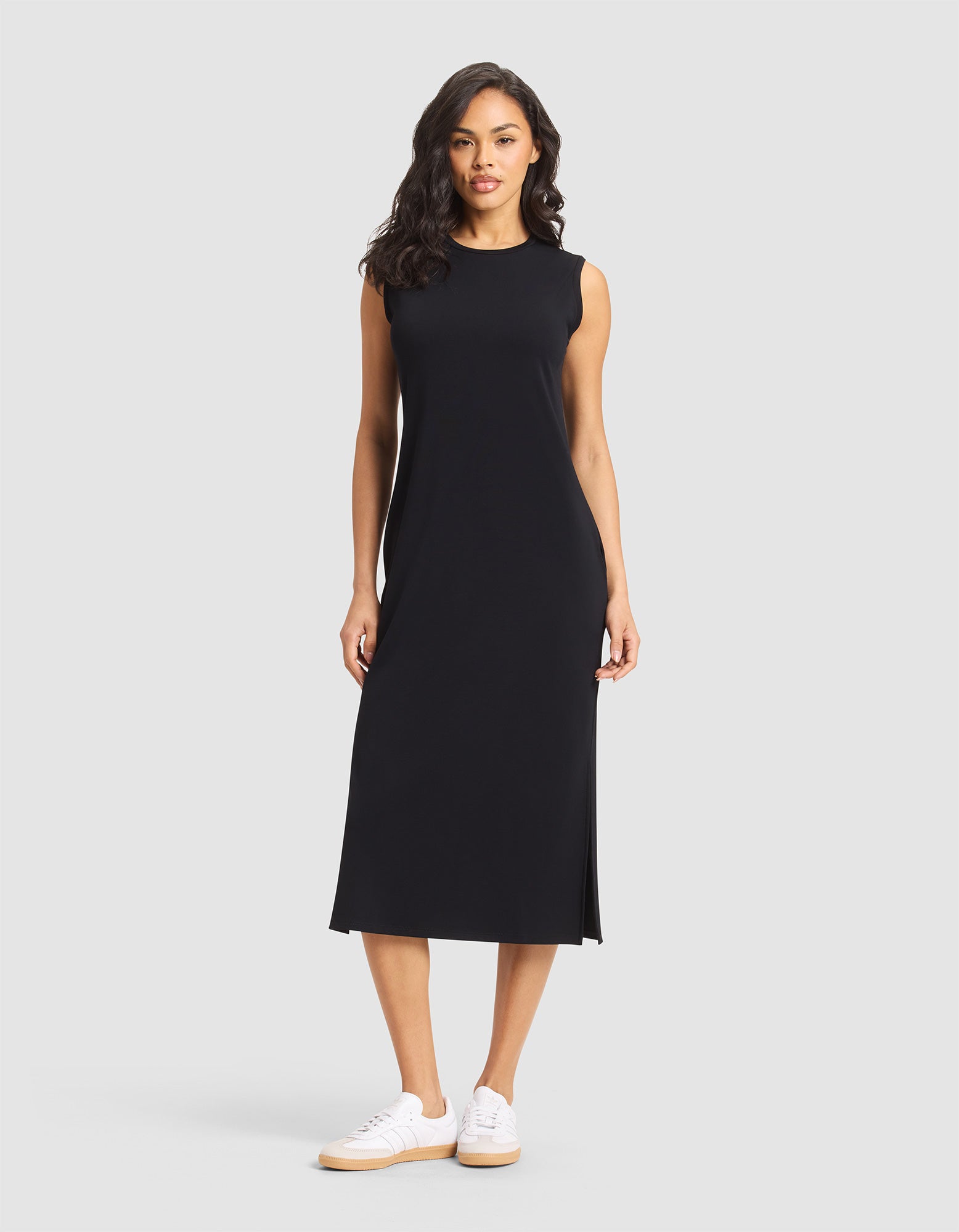 A woman with long, wavy hair and a neutral expression stands against a light gray background wearing the Cozy Earth Women's Brushed Bamboo Sleeveless Midi Dress in black, featuring side slits, paired with white sneakers. |Color:Black