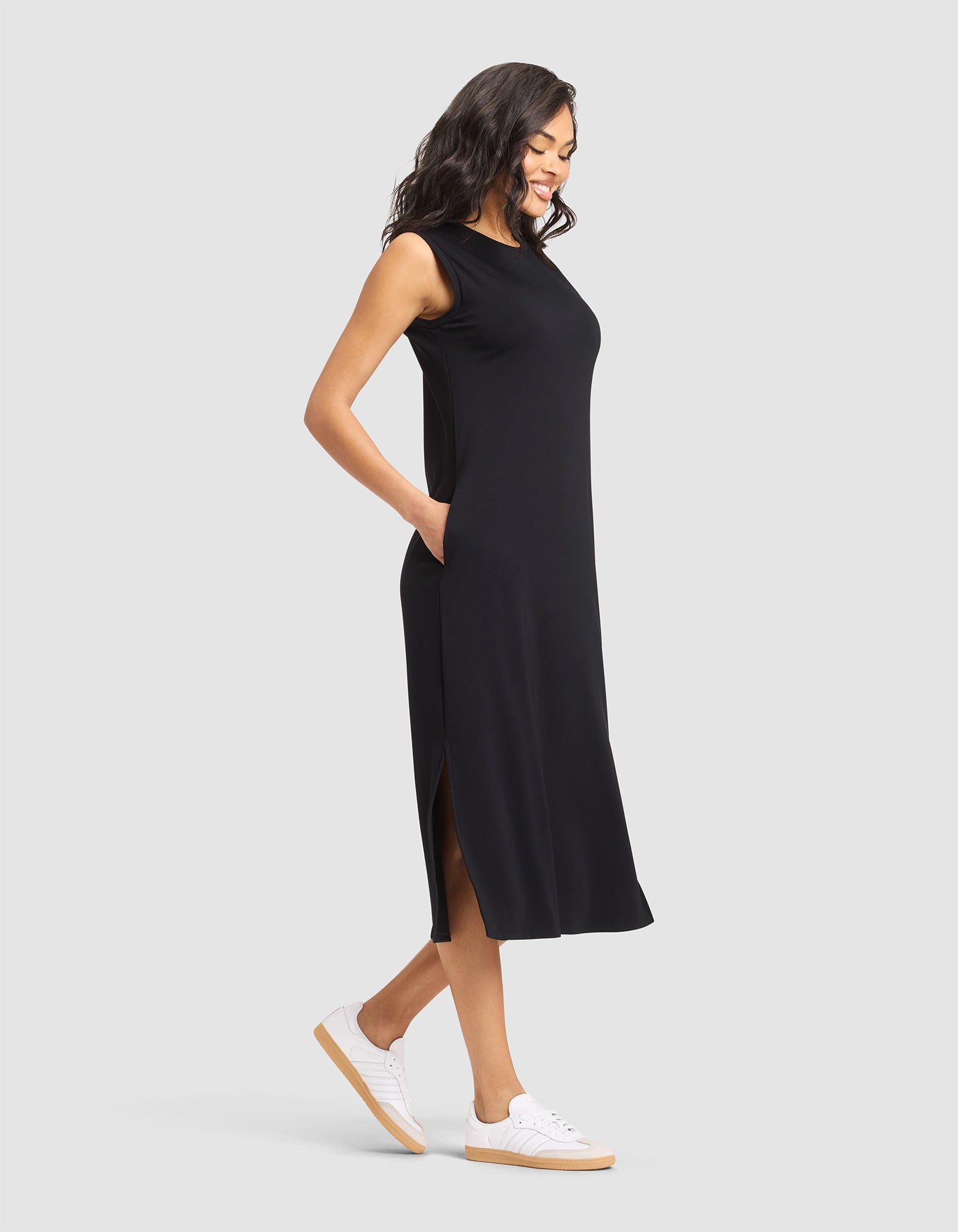 A woman smiles with her hands in her pockets, wearing the Cozy Earth Women's Brushed Bamboo Sleeveless Midi Dress and white sneakers, standing against a plain light gray background. |Color:Black