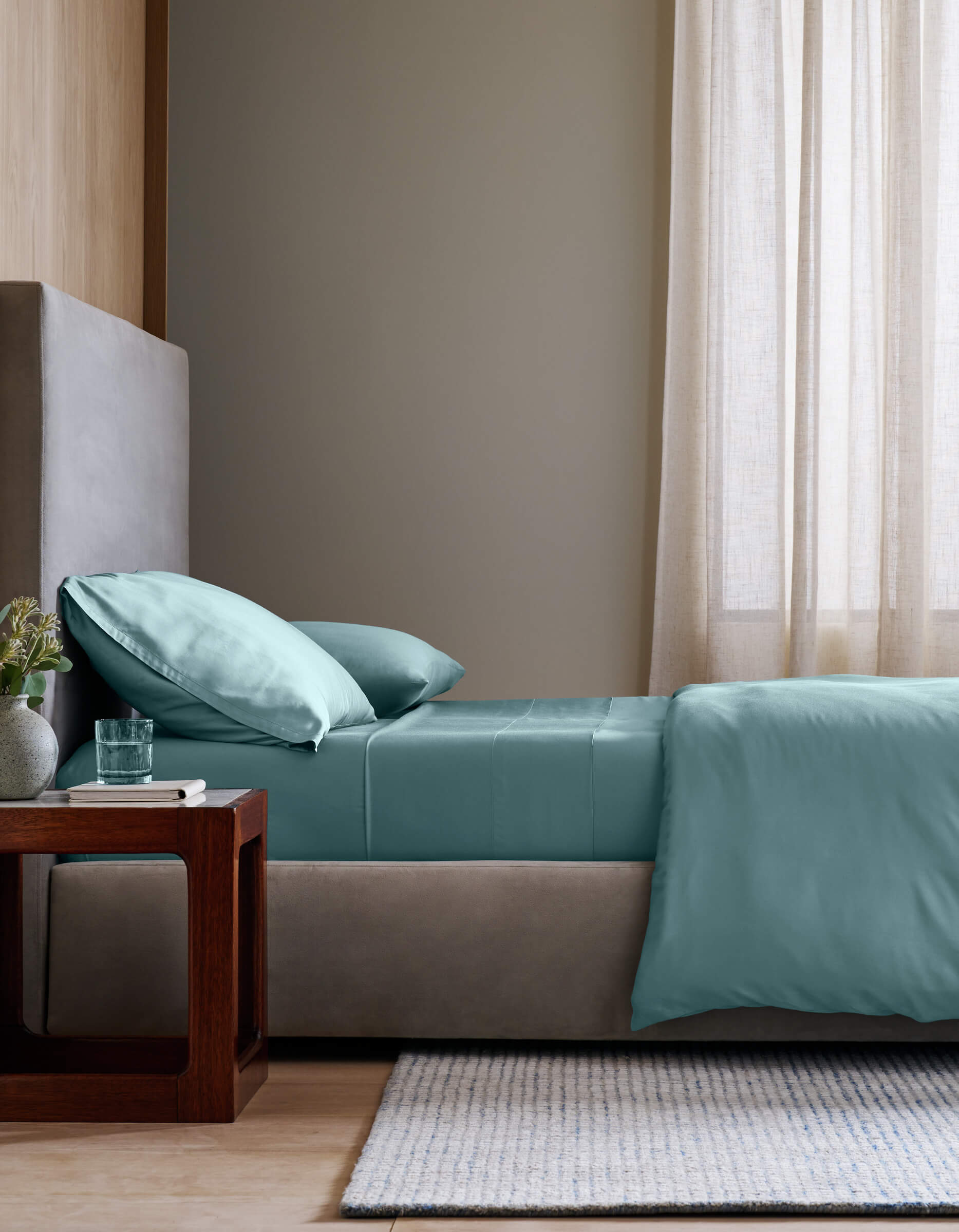 A modern bedroom features a neatly made bed with Cozy Earth's Bamboo Sheet Set in teal and two pillows, next to a wooden nightstand holding a glass of water and a small potted plant. Soft natural light filters through sheer beige curtains.|Color:River