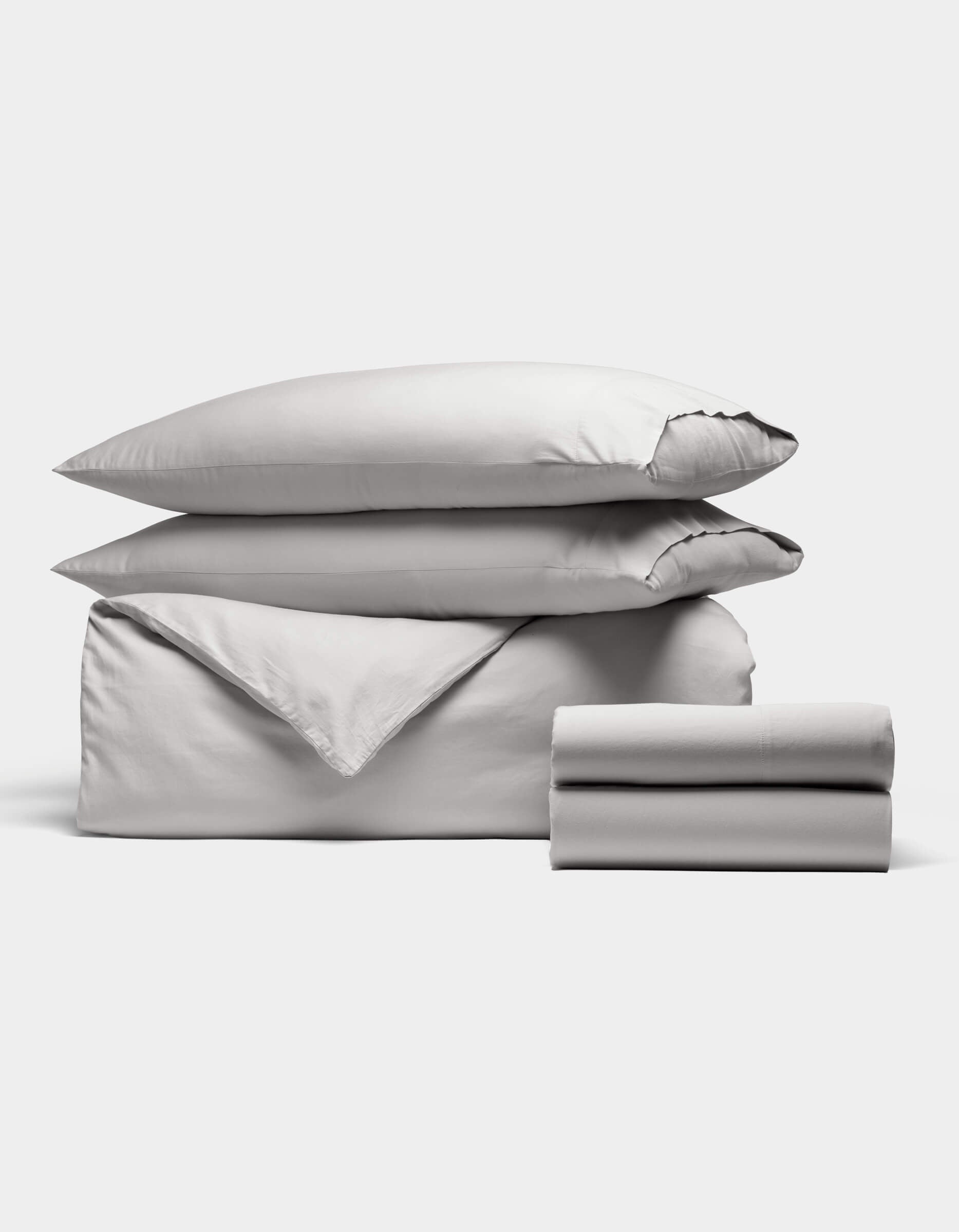 The Soft-Wash Cotton Bedding Deluxe Bundle by Cozy Earth features a neatly stacked set of light gray bedding—two pillows, a folded duvet, and two folded sheets—arranged against a plain white background.|Color:Dew