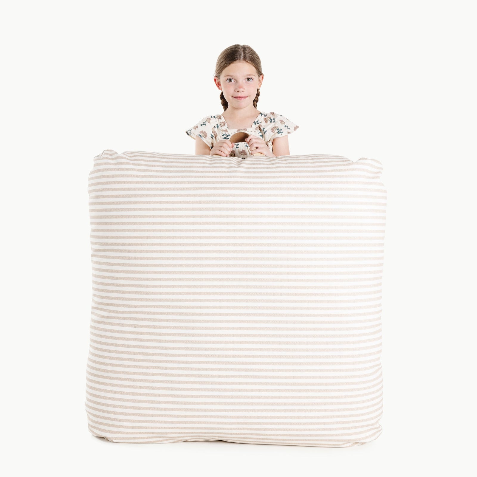 A young girl with braided hair stands behind the large Gathre Wholesale Floor Cushion in Cafe Stripe, holding the top edge and smiling at the camera against a plain white background.