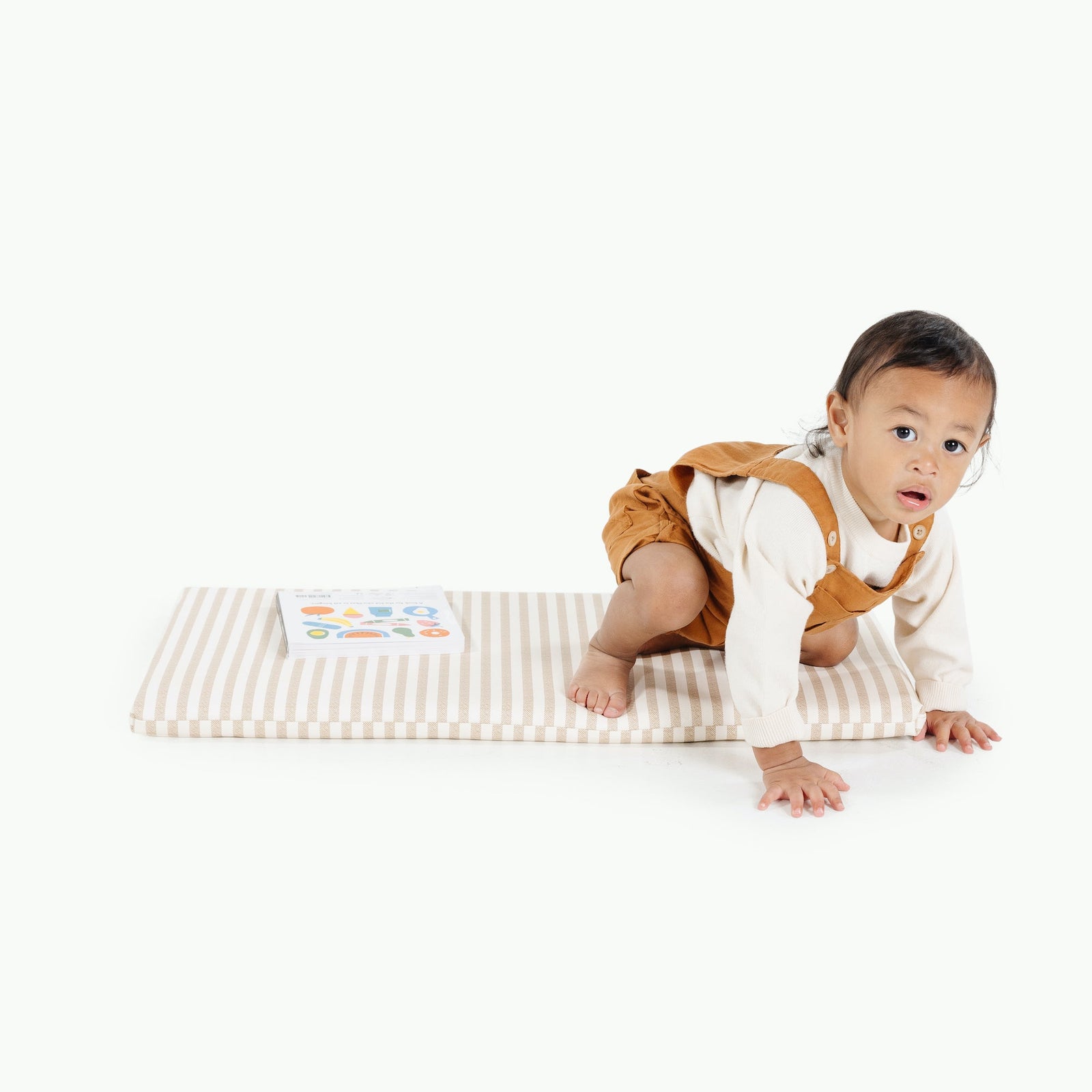 A young child in a cream shirt and brown overalls crawls on the Gathre Wholesale Padded Changing Mat in Cafe Stripe, with a book beside them, set against a white background.