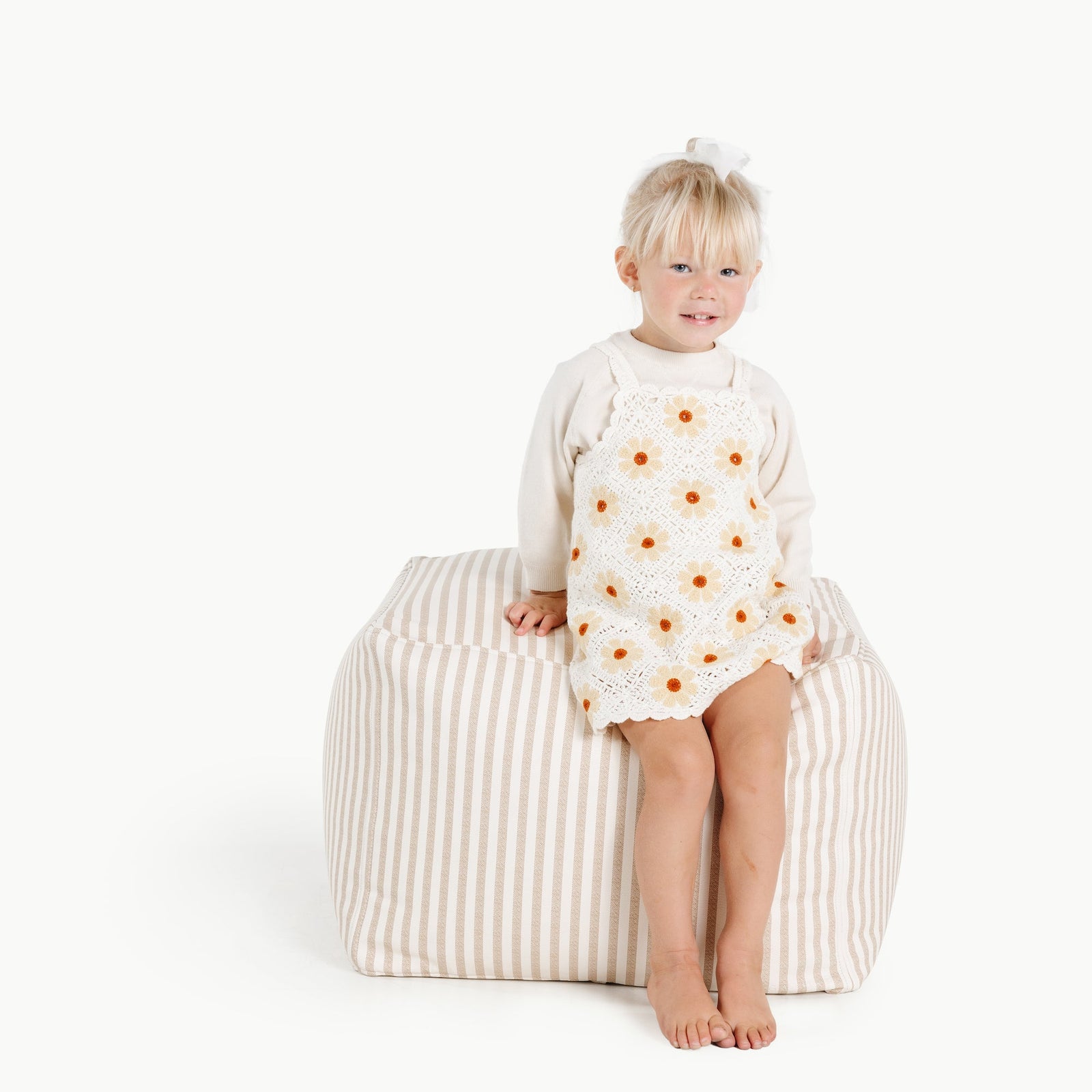 A young child sits barefoot on a Gathre Wholesale Pouf in Cafe Stripe, wearing a cream long-sleeve shirt and a white dress with orange flowers. The background is plain white.