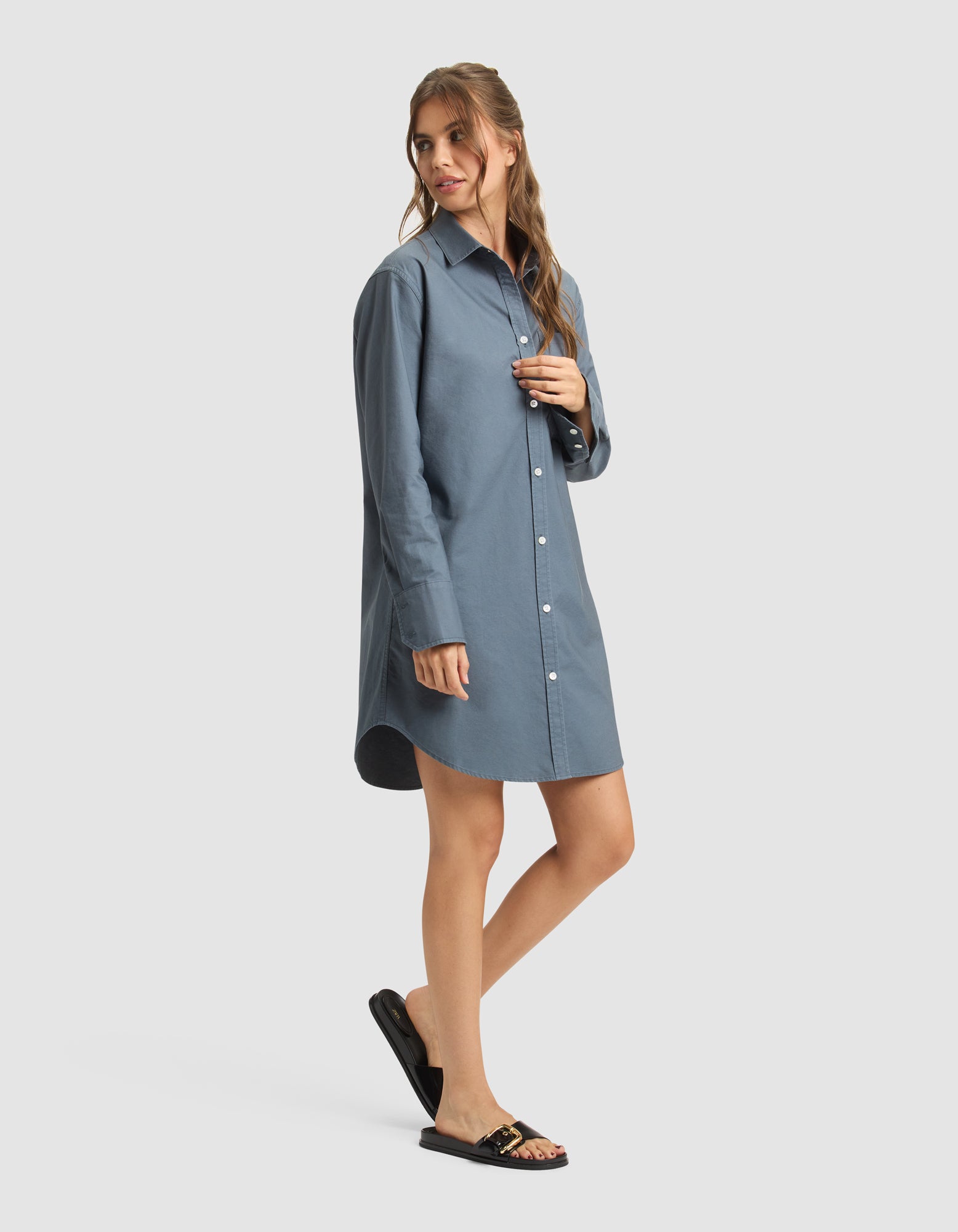 A woman poses in a relaxed way, wearing Cozy Earth's Women's Camden Dress—a loose-fitting, long-sleeve blue shirt dress—paired with black sandals featuring gold accents against a plain light background.