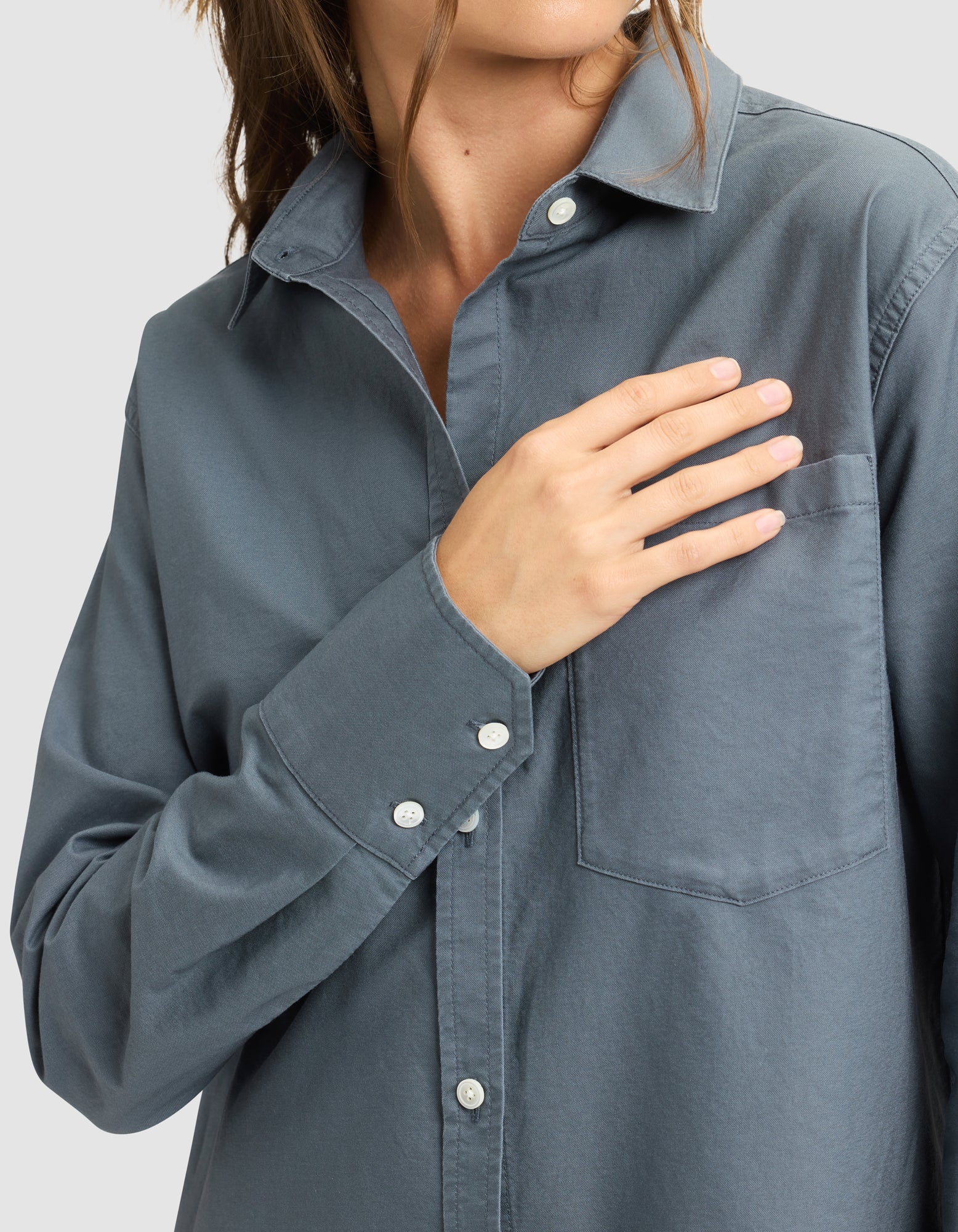 A woman wearing the Cozy Earth Women's Camden Dress touches the chest pocket with her right hand. Her face is partially out of frame, and the background is plain light gray.