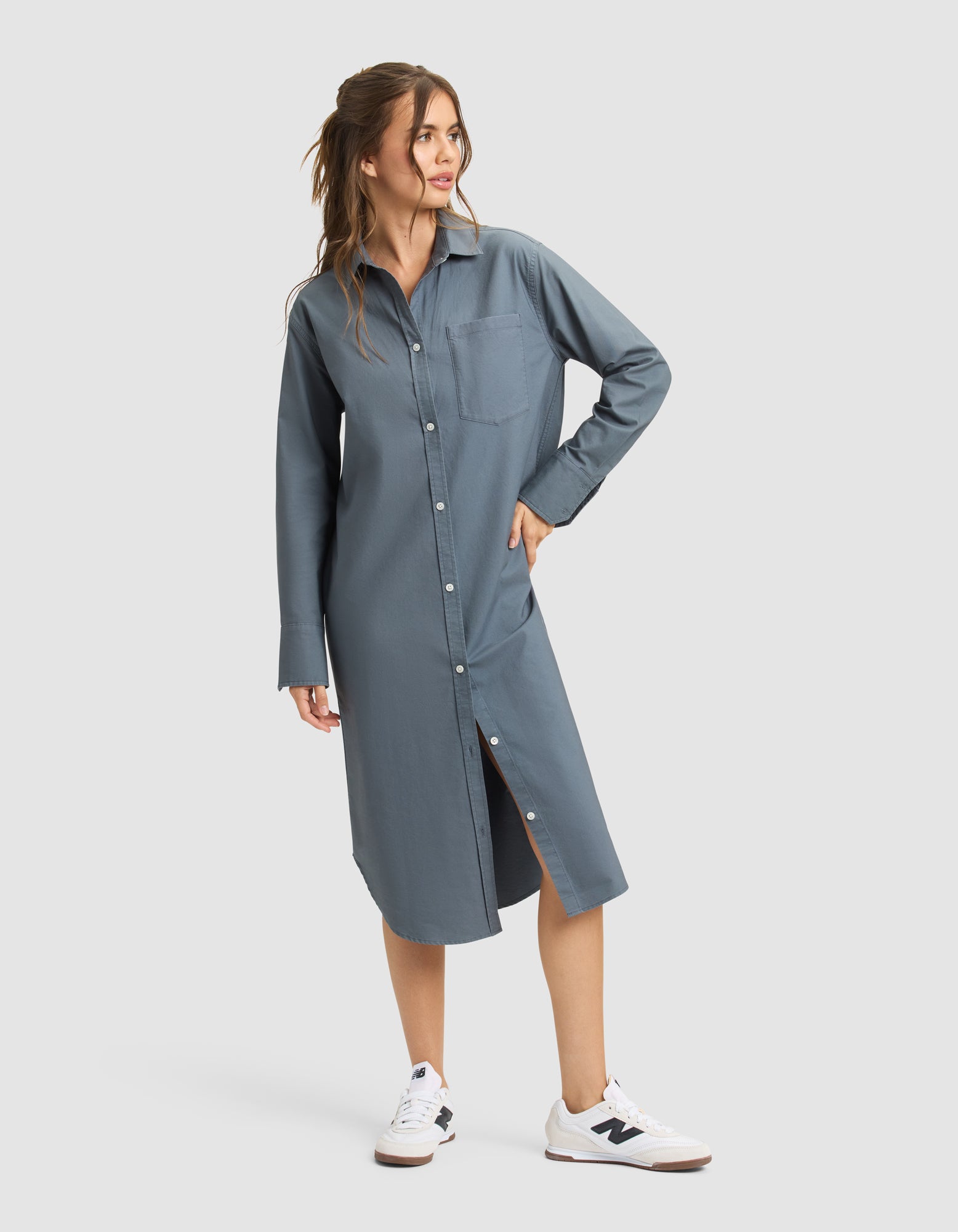 A woman with long, wavy brown hair models the Cozy Earth Women's Camden Midi Dress—a blue-gray, long-sleeve button-down shirt dress with a chest pocket—styled with white sneakers against a plain light background.