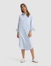 A woman wears the Cozy Earth Women's Camden Midi Dress, a loose light blue, long-sleeve, button-down shirt dress with subtle stripes. She pairs it with white sneakers and smiles slightly, posing with one hand by her side. |Color:Blue Oxford Stripe