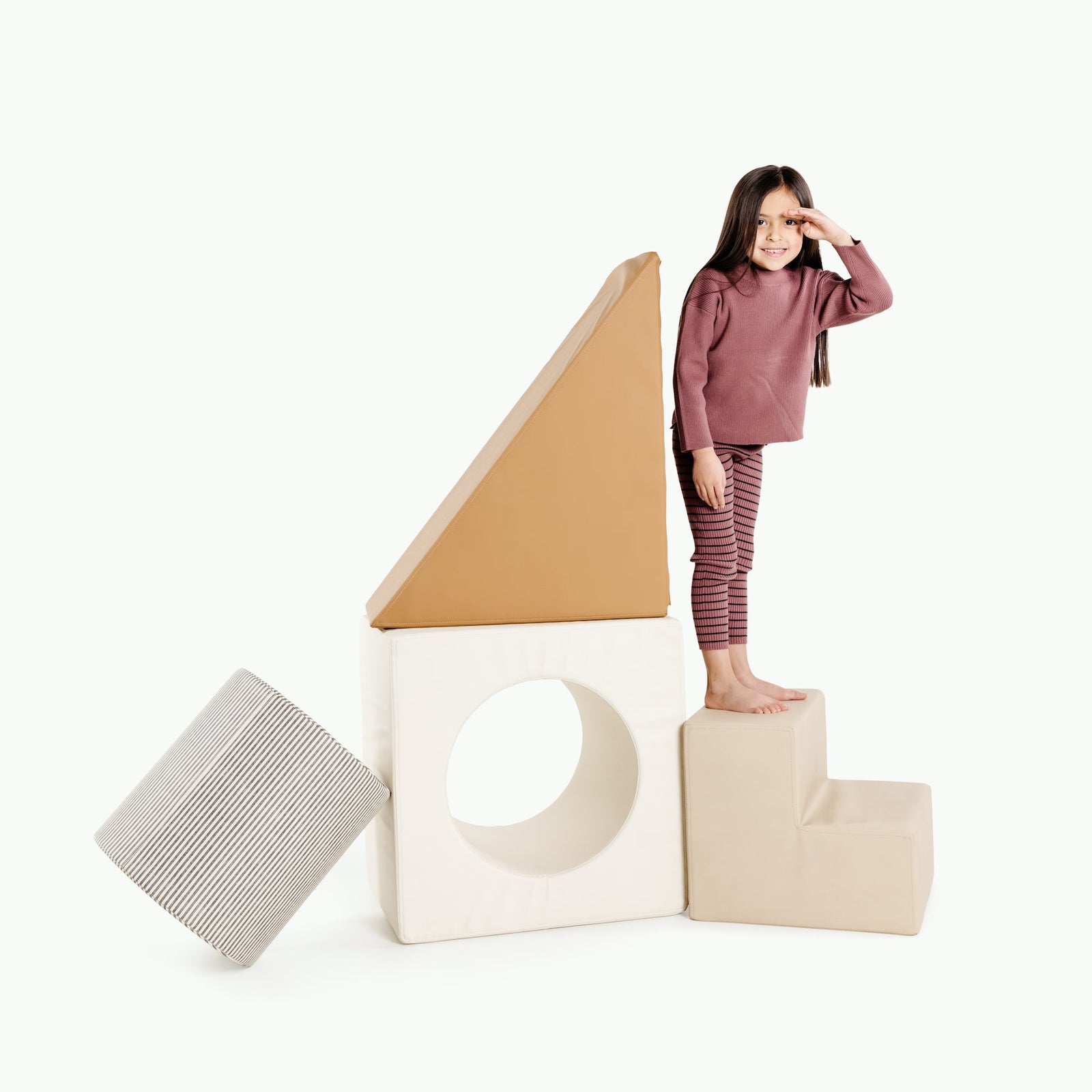 A young girl smiles and shades her eyes while standing on Gathre's Wholesale Block Playset, featuring beige foam blocks and large soft shapes—a cylinder and triangle—arranged like a house.