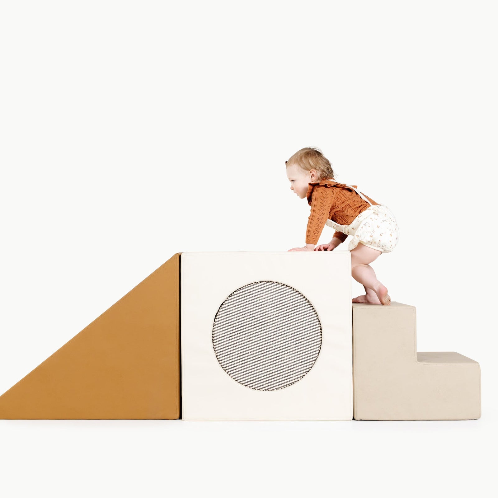 A young child climbs over Gathre's Wholesale Block Playset, featuring soft, foam stair and ramp blocks in beige, brown, and white, with a circular design on one block, shown on a plain white background.