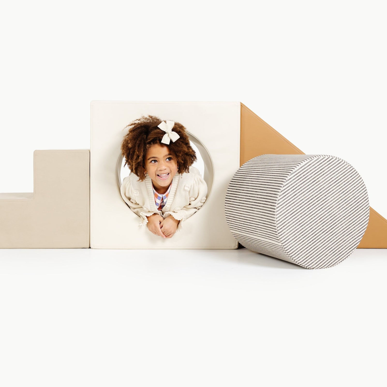 A young girl with curly hair and a white bow smiles while crawling through the Gathre Wholesale Block Playset, surrounded by geometric foam shapes on a white background.