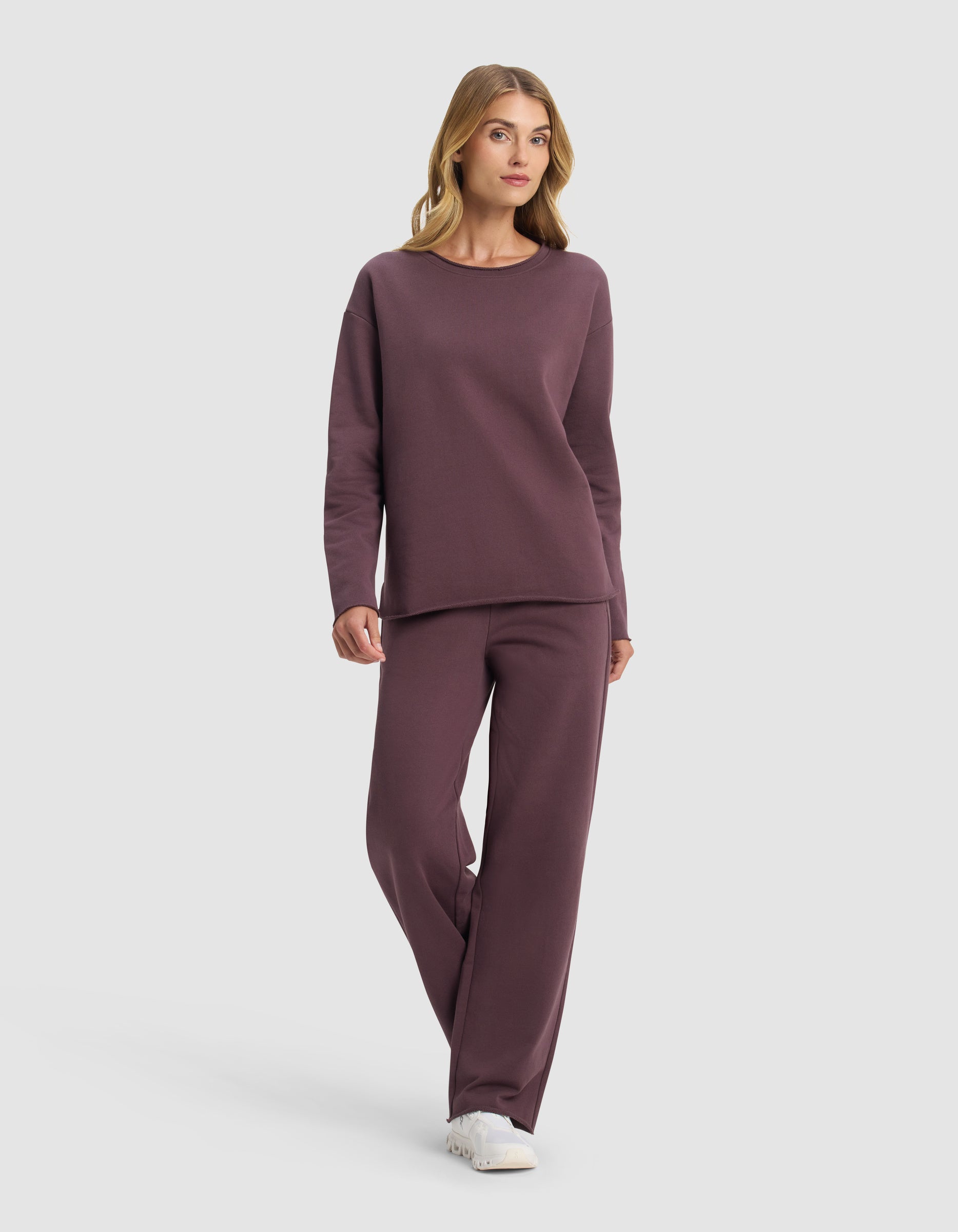 A woman with light brown hair models the Cozy Earth Women's Chelsea Set— a loose-fitting, long-sleeve mauve sweatshirt with matching pants—paired with white sneakers, against a plain white background.|Color:Fig
