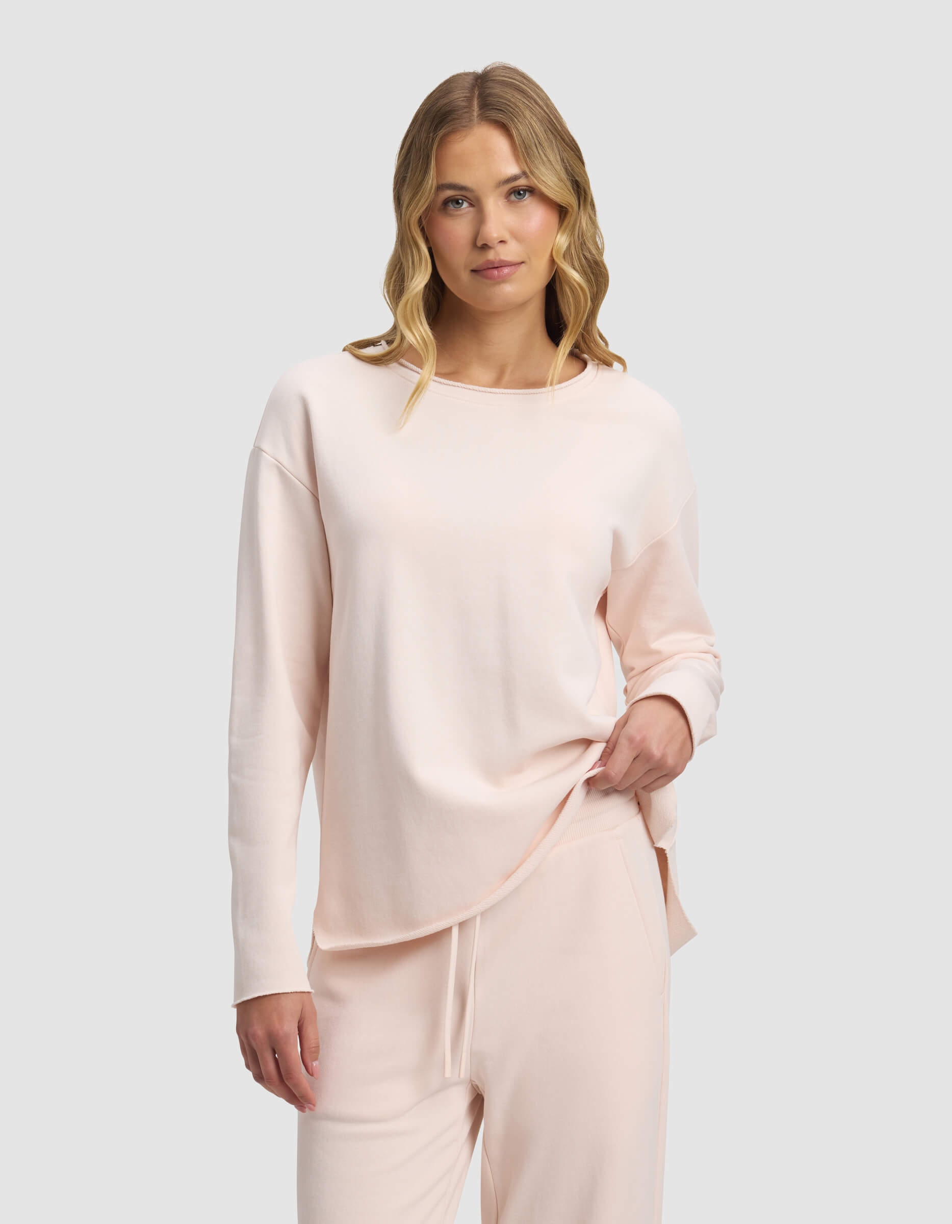 A woman with wavy blonde hair wears the Cozy Earth Women's Chelsea Pullover in light pink and matching drawstring pants, standing against a plain light gray background while looking at the camera and holding part of her shirt hem.|Color:Petal