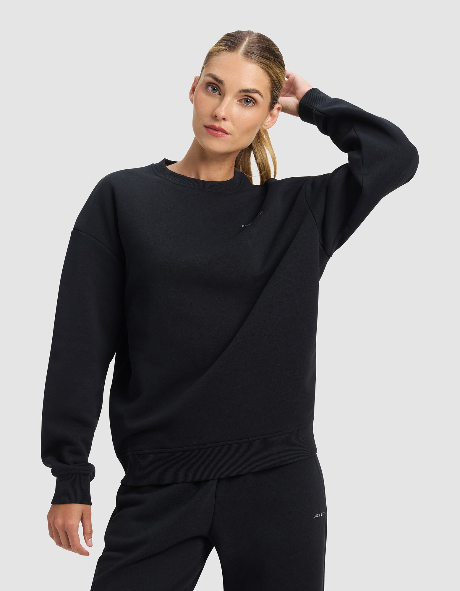 A woman wears the Cozy Earth Women's Cityscape 2.0 Crewneck in black, paired with matching pants, standing against a light gray background with one hand behind her head and a neutral expression.|Color:Jet Black
