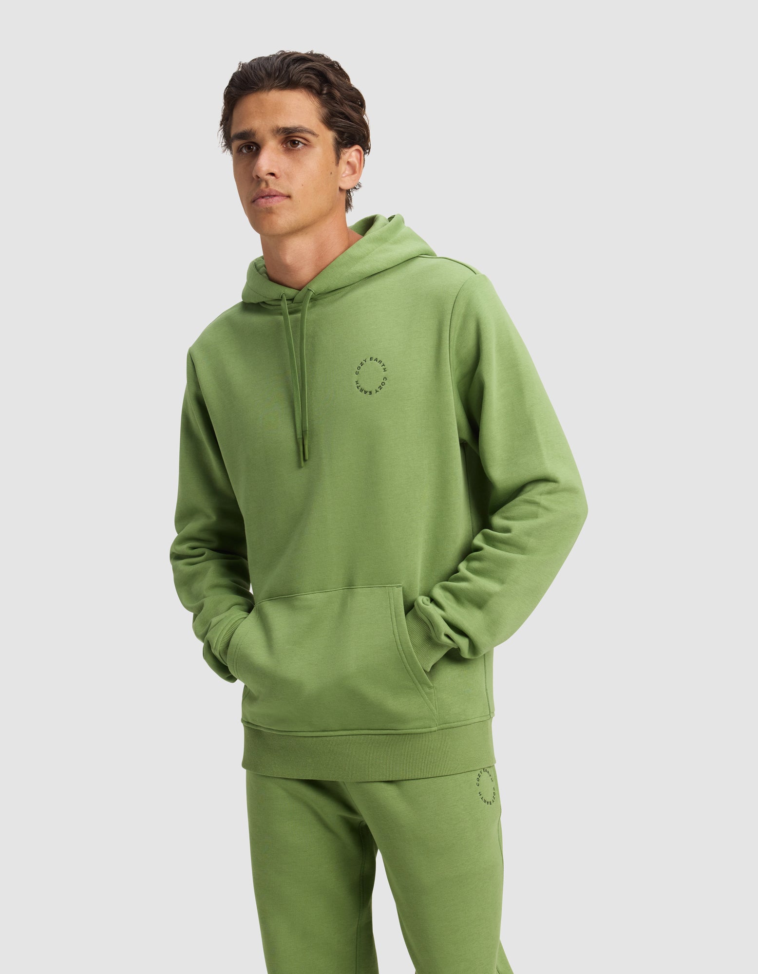 A man wears a Cozy Earth Men's Cityscape 2.0 Hoodie and matching sweatpants in Clover, both featuring a small circular stitched design, with hands in the hoodie pocket against a plain light gray background.|Color:Clover