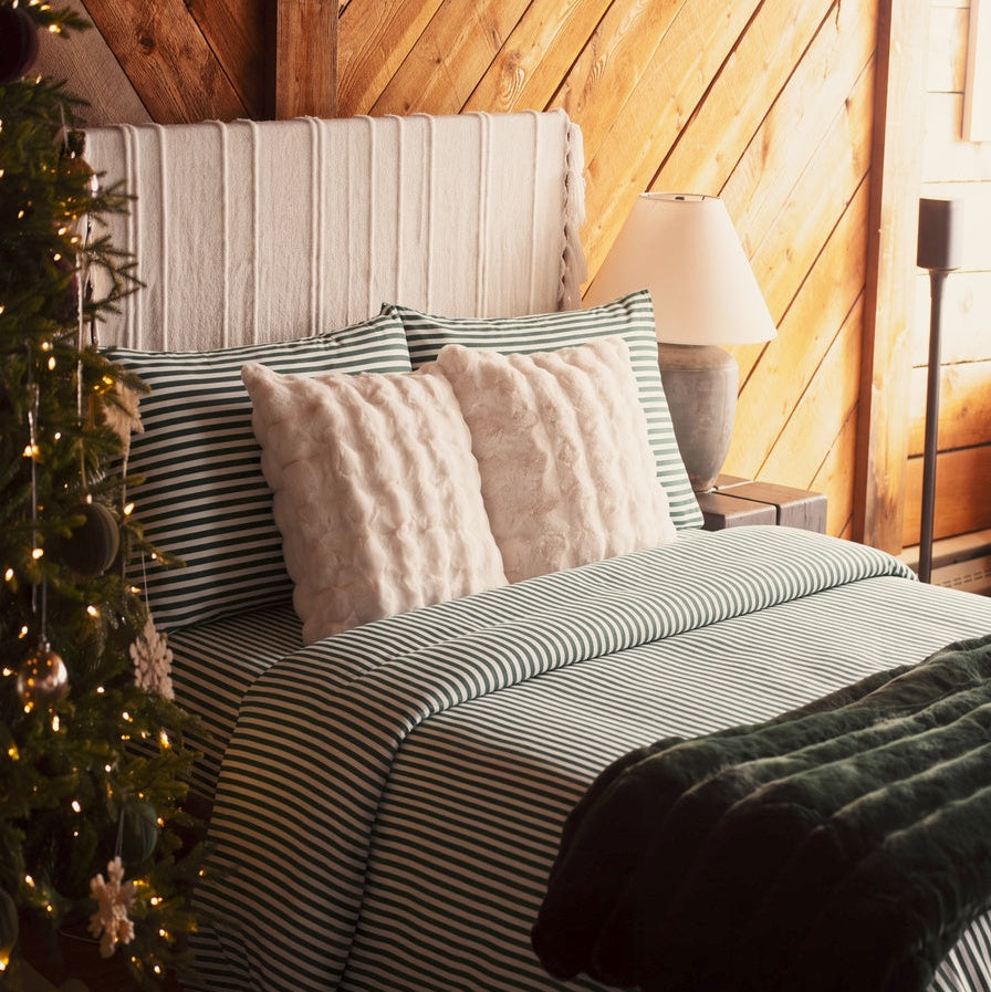 A cozy bedroom scene with a bed dressed in striped bedding, two textured cream pillows, a wooden slat headboard, warm wood-paneled walls, a bedside lamp, and a decorated Christmas tree at the side.
