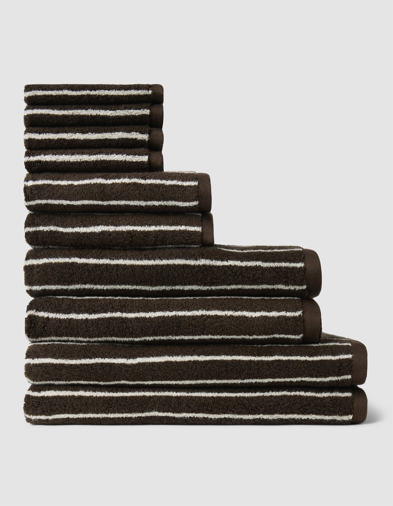 The Complete Striped Luxe Bath Bundle by Cozy Earth features six brown towels with white stripes, neatly stacked in descending size against a plain light background.|Color:Espresso Coconut Stripe
