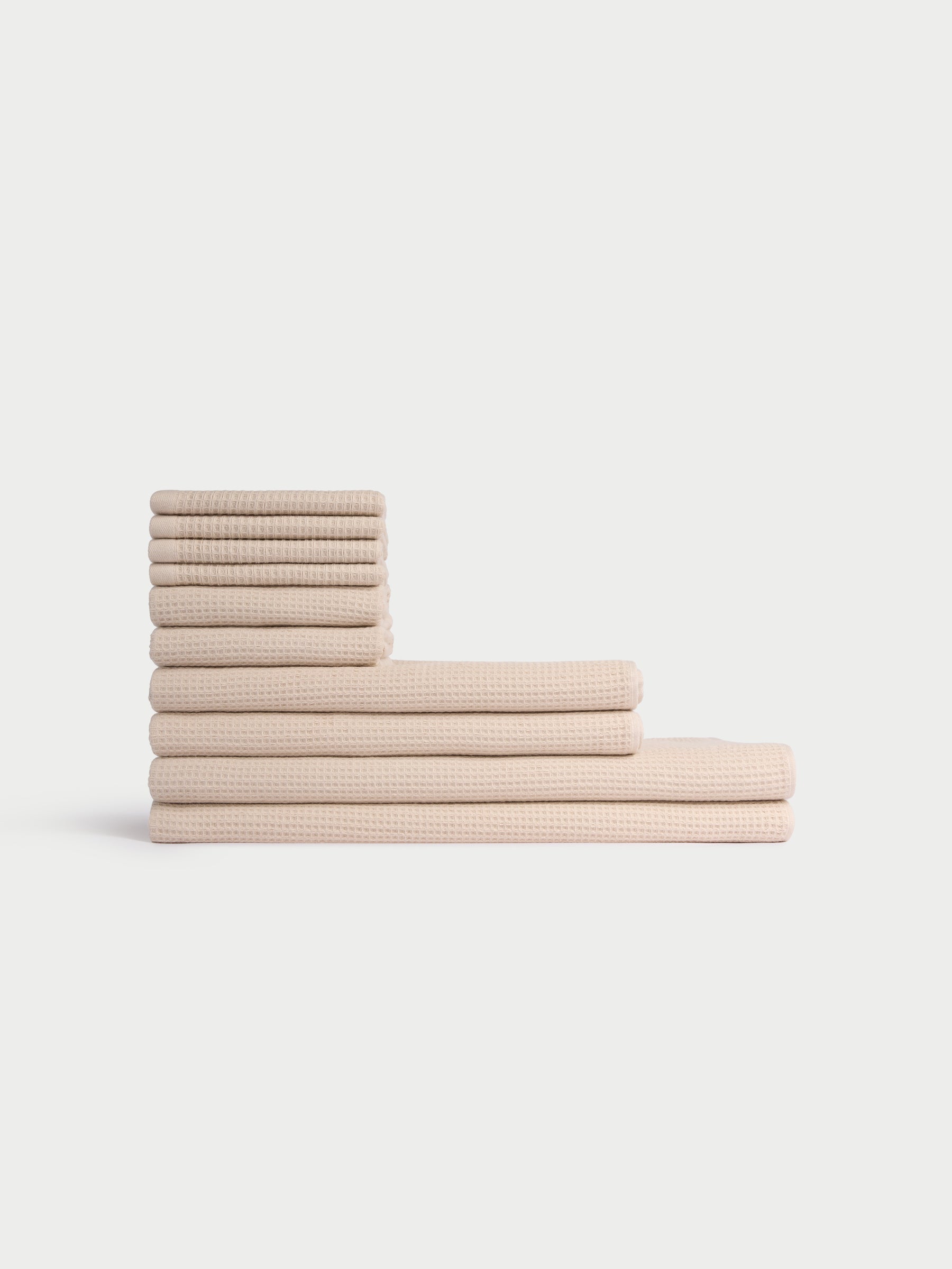 The Cozy Earth Complete Waffle Bath Bundle features beige, waffle-textured towels neatly stacked in two rows on a white background, with smaller towels stacked vertically and larger ones laid horizontally at the bottom. |Color:Birch