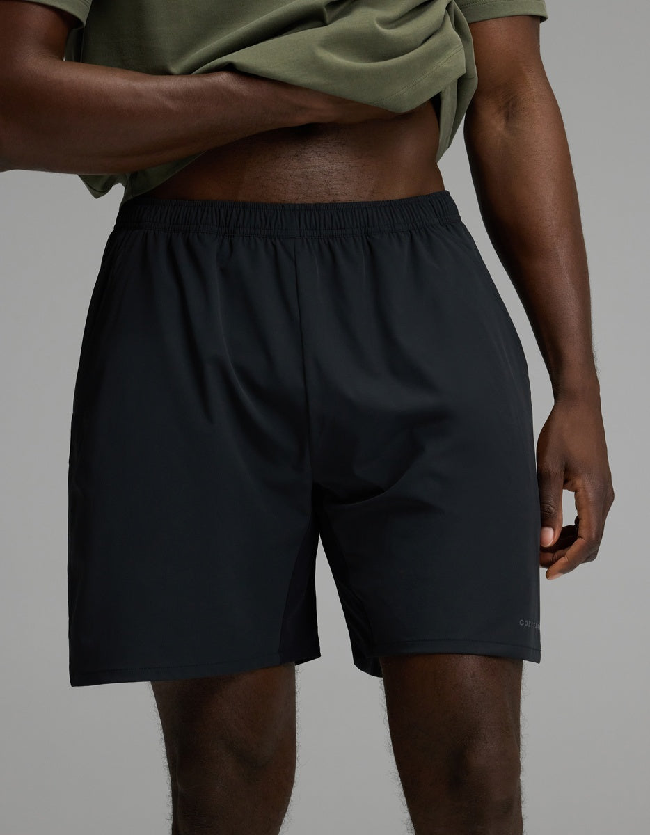A person wearing a green shirt and the Cozy Earth Men’s Day & Night Short 7″ in Jet Black stands with one hand on their hip against a plain gray background.|Color:Jet Black