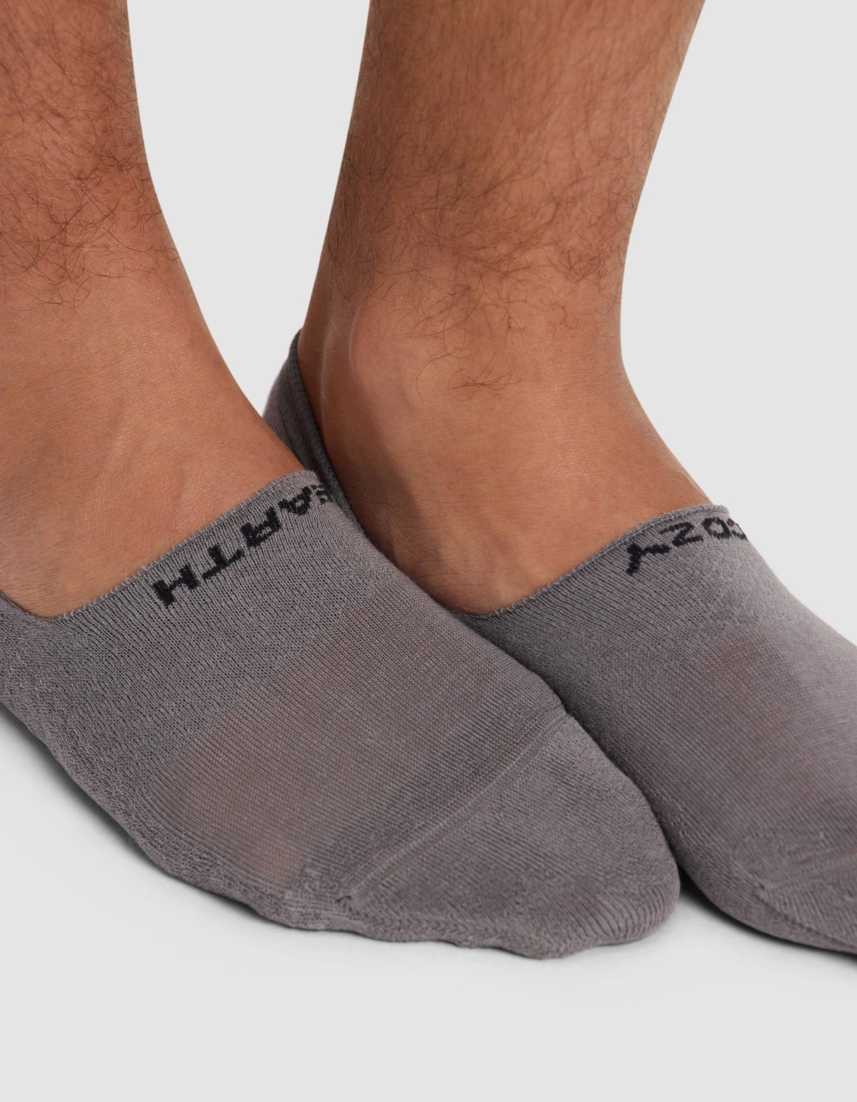 Close-up of a person's feet wearing Cozy Earth Essential No-Show Socks in gray with black text near the cuffs, standing on a white background.