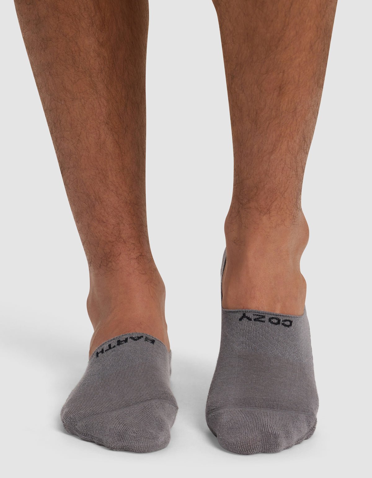 A close-up shows a person's lower legs and feet on a white surface, wearing Cozy Earth's Essential No-Show Socks in gray, each featuring black text on top.