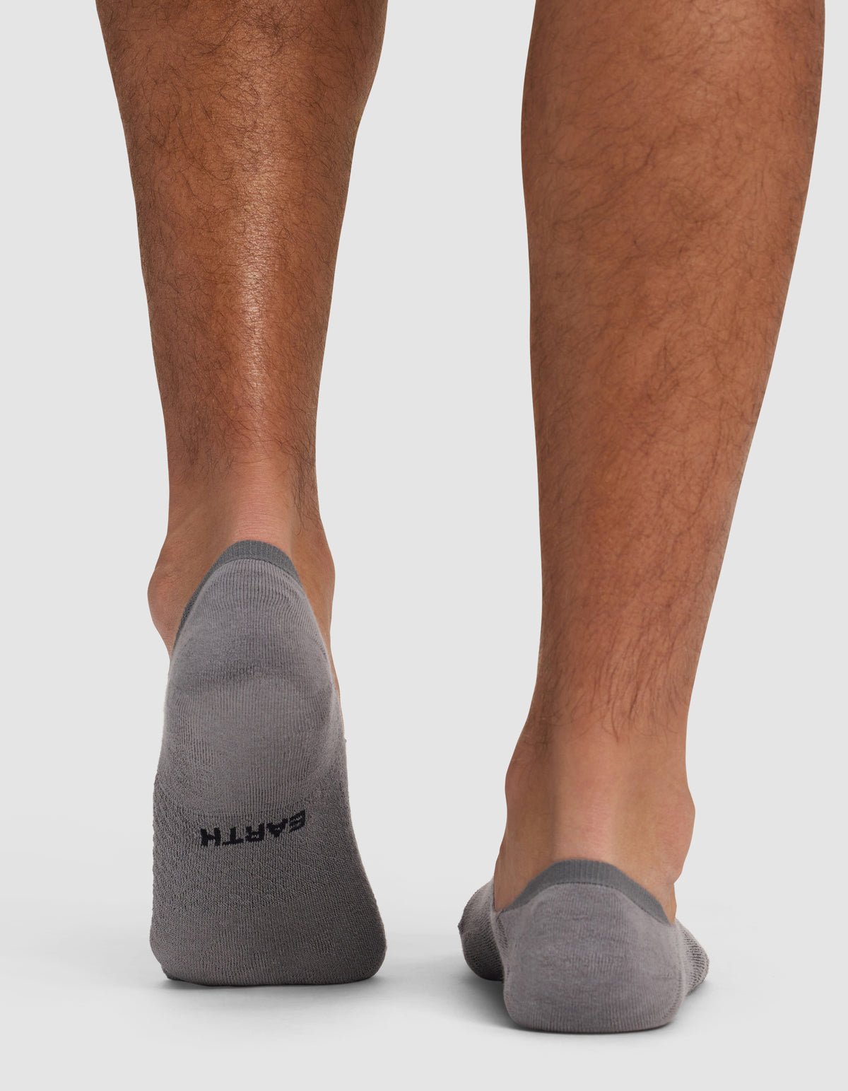 A close-up shows the back of a person's lower legs and feet in gray Cozy Earth Essential No-Show Socks. "EARTH" is printed in black on the left sock. The background is plain white.
