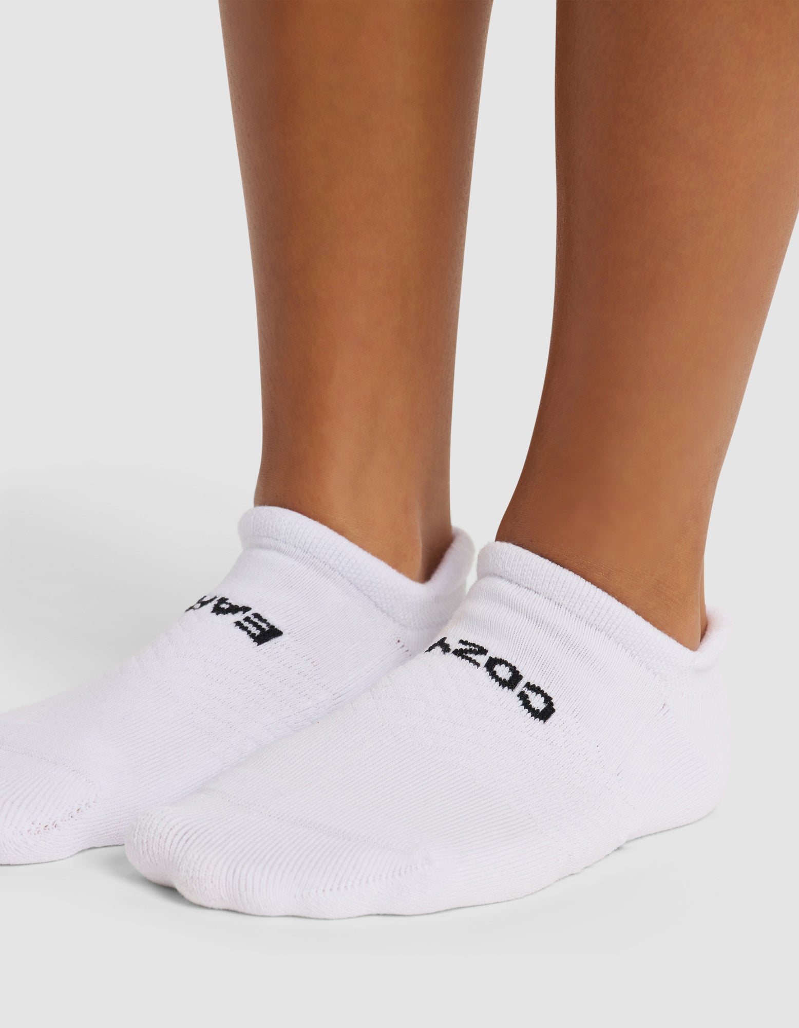 Close-up of a person wearing Cozy Earth Essential Ankle Sock 4-Pack in white with black text, standing on a light surface. Only their lower legs and feet are shown.