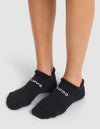 A person wearing Cozy Earth's Essential Ankle Socks stands on a light gray surface. The black socks feature white "COZY" text on the right foot and "EARTH" on the left. |Color:Black