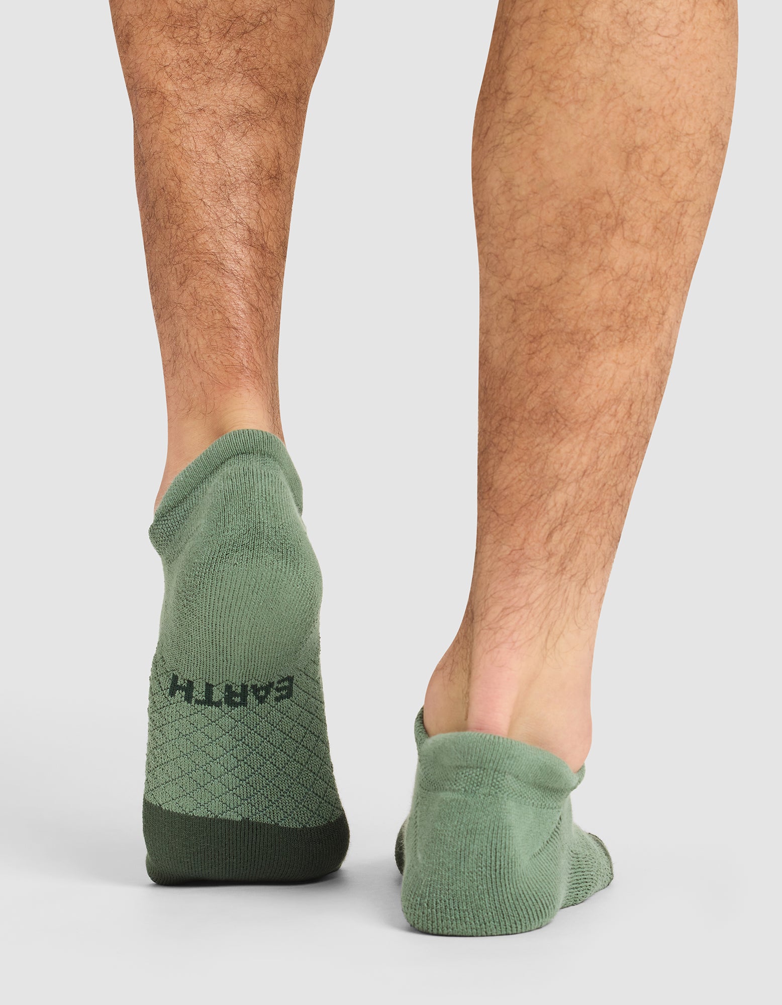 A person stands on a white background wearing Cozy Earth Essential Ankle Socks in green; the left sock fits properly, while the right one is slipped off the heel and bunching underfoot.