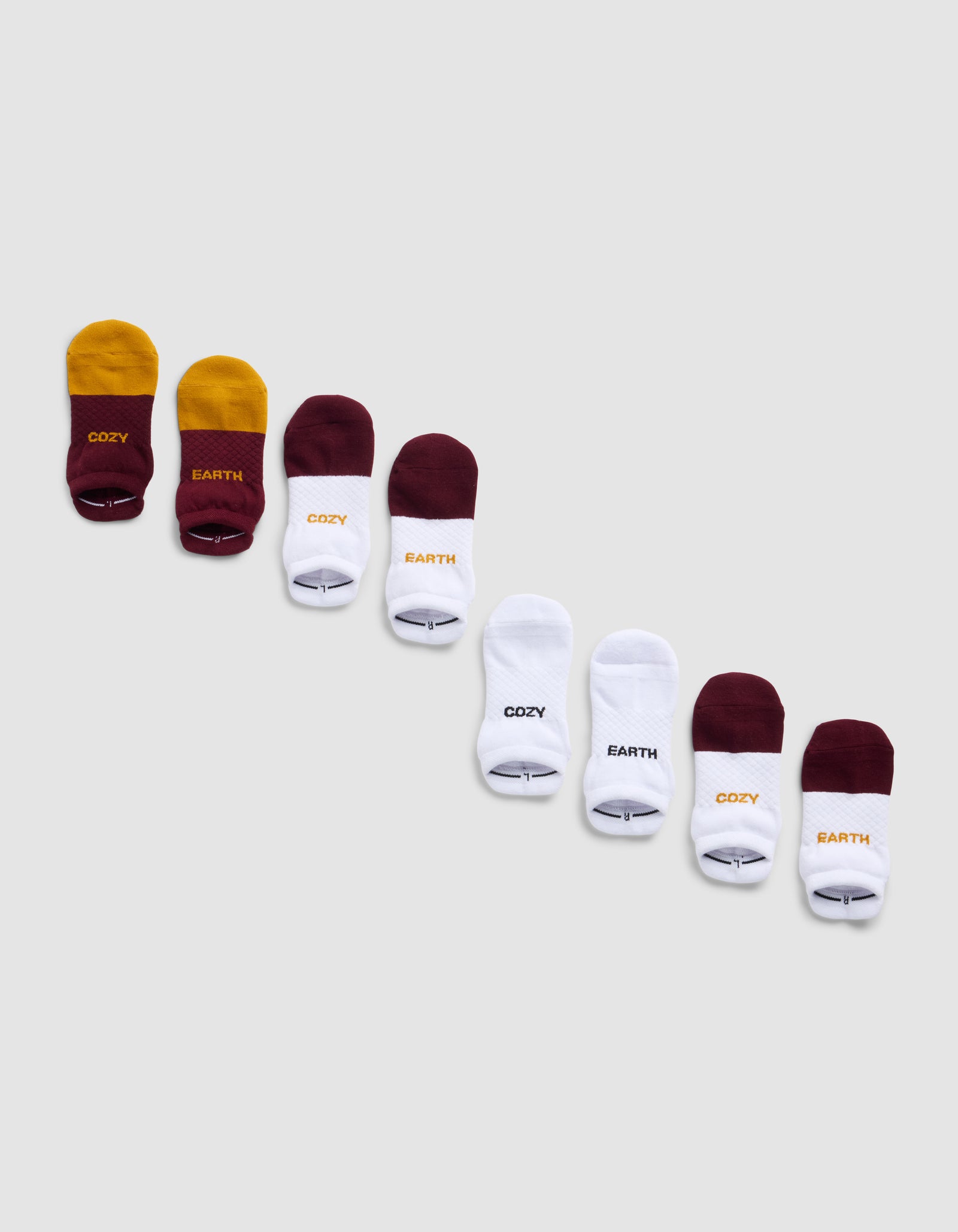 The Cozy Earth Essential Ankle Sock 4-Pack features socks in yellow, burgundy, and white arranged diagonally on a light gray background. Some socks have "COZY" or "EARTH" printed near the toes.