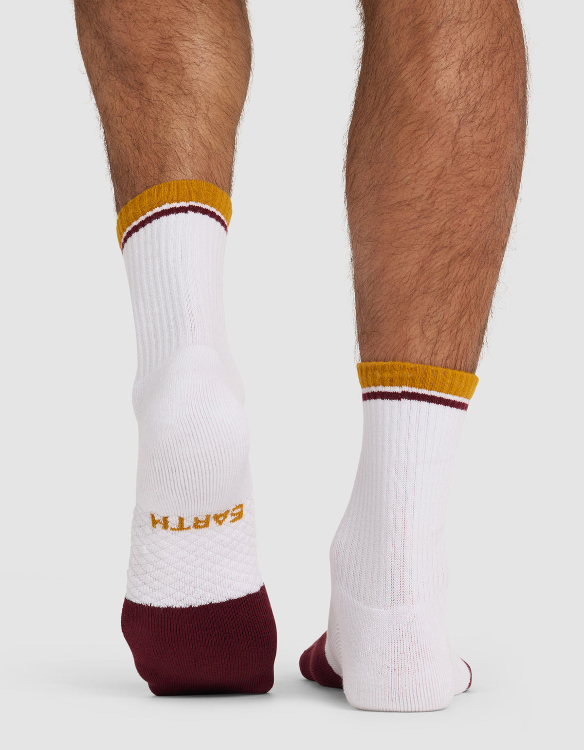 A person wears Cozy Earth Essential Calf Socks—white with gold "EARTH" on the sole, maroon heel and toe, and gold/maroon accents—standing on a white surface.