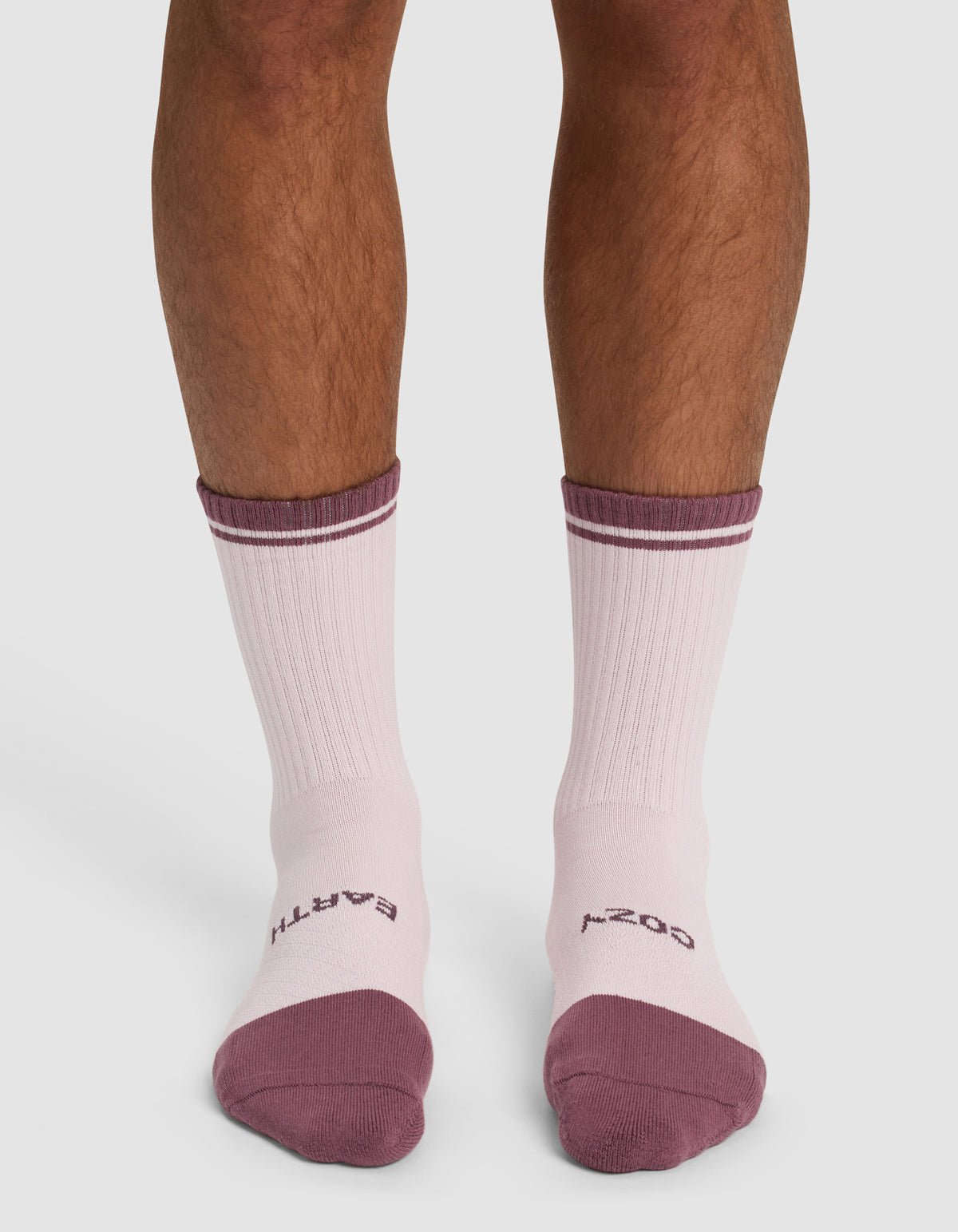 A pair of legs wears Cozy Earth Essential Calf Socks in light pink with purple toes, heels, and top stripes. “EARTH” and “COZY” are printed in purple on the sock tops.