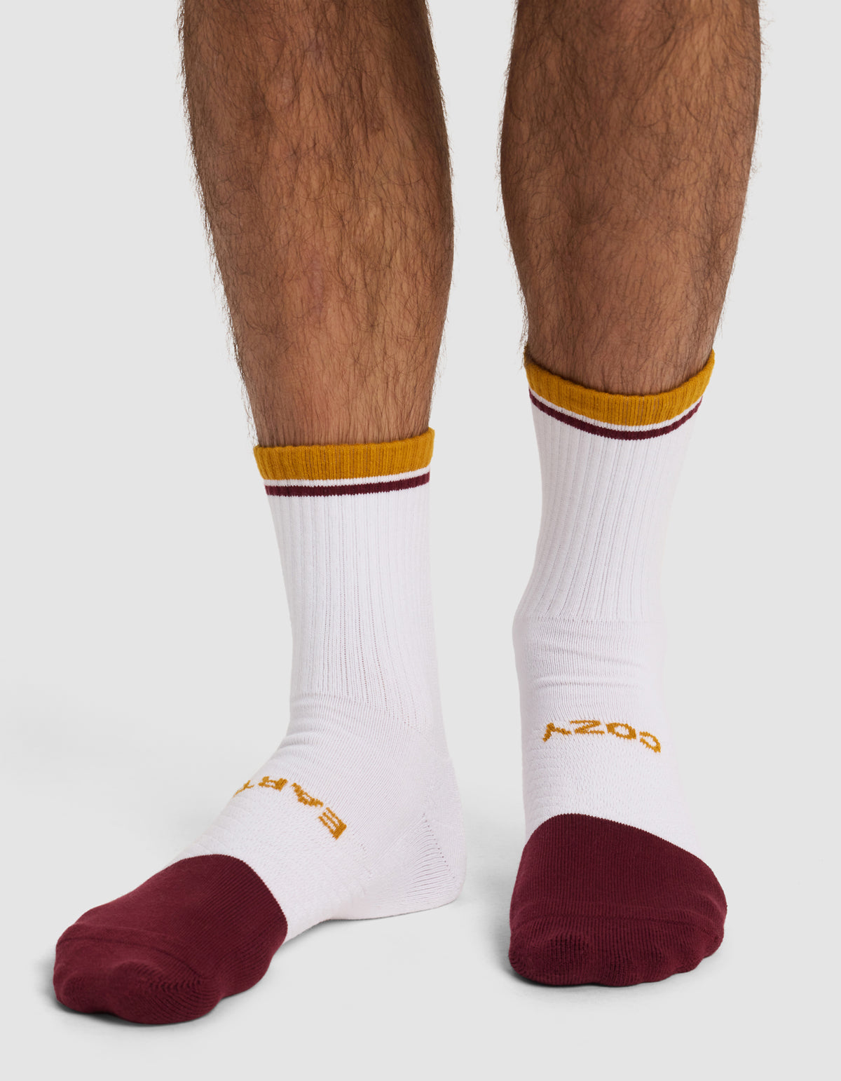 A person wears Cozy Earth Essential Calf Socks (from the 4-Pack), featuring white fabric with gold and maroon stripes at the top, maroon toes, and gold text on the upper foot. Only their lower legs are visible.