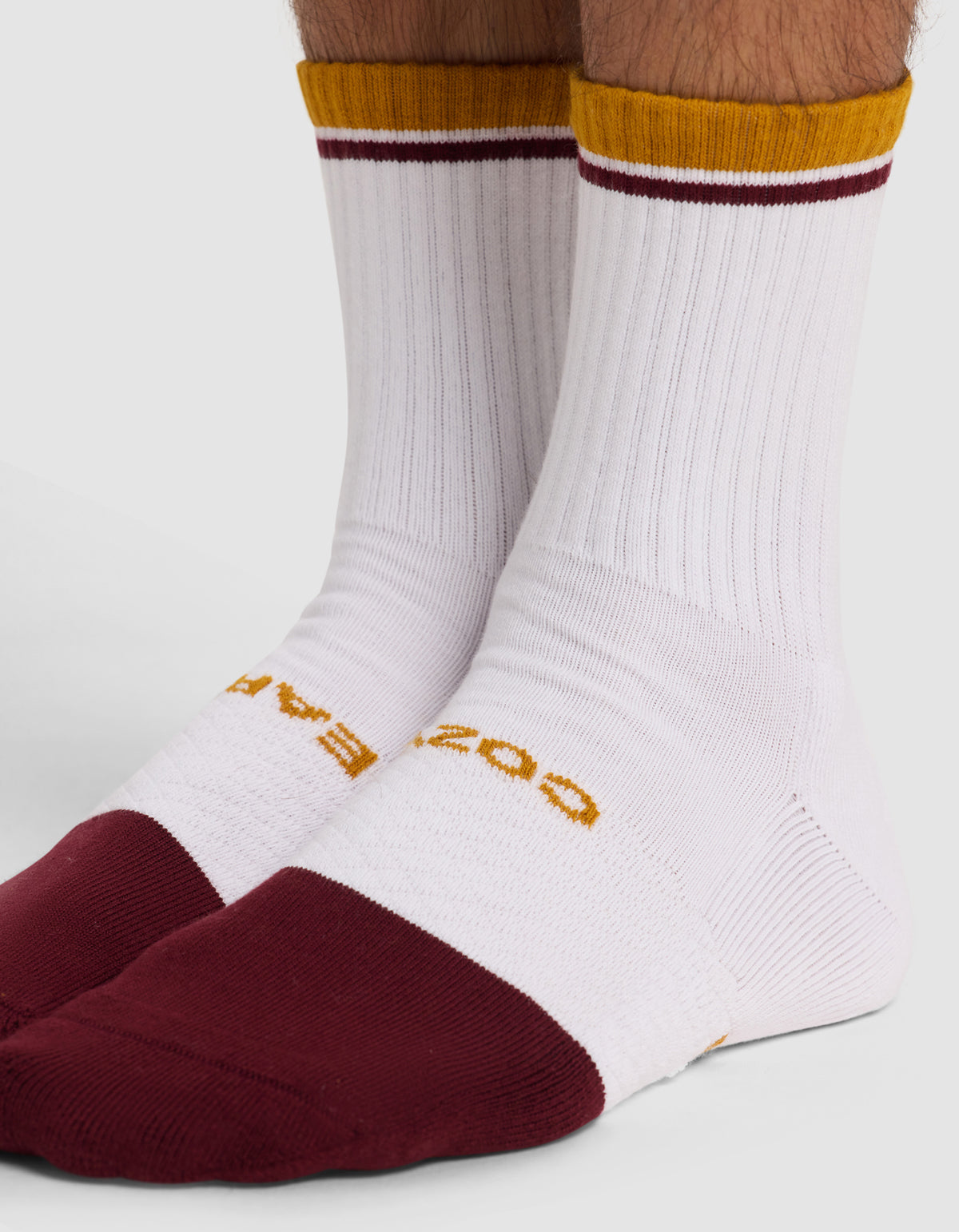 A close-up of a person wearing Cozy Earth's Essential Calf Sock, a white ribbed mid-calf sock with maroon toes and heels, a gold top stripe, and yellow text by the toes, shown as part of the 4-pack set.