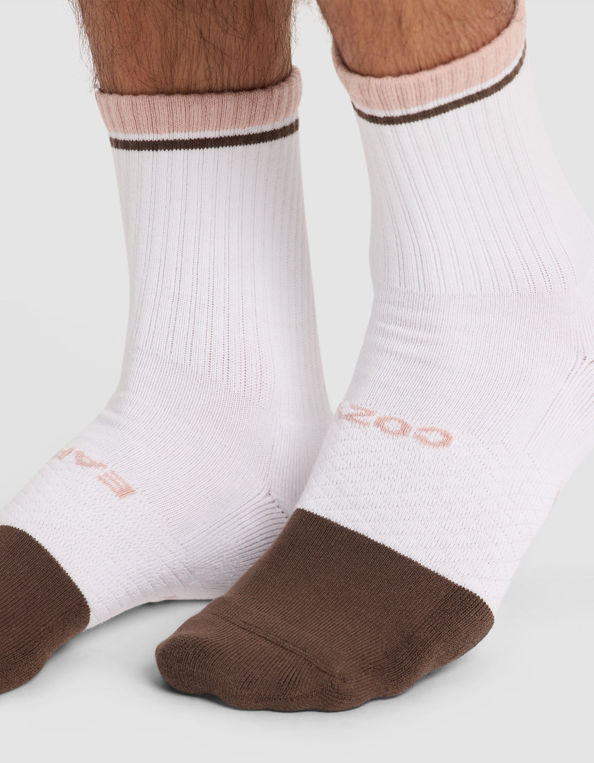 Two feet wear Cozy Earth Essential Calf Socks—white ribbed socks with brown toes and soles, pink accents at the top, a thin brown ankle stripe, and subtle pink text.
