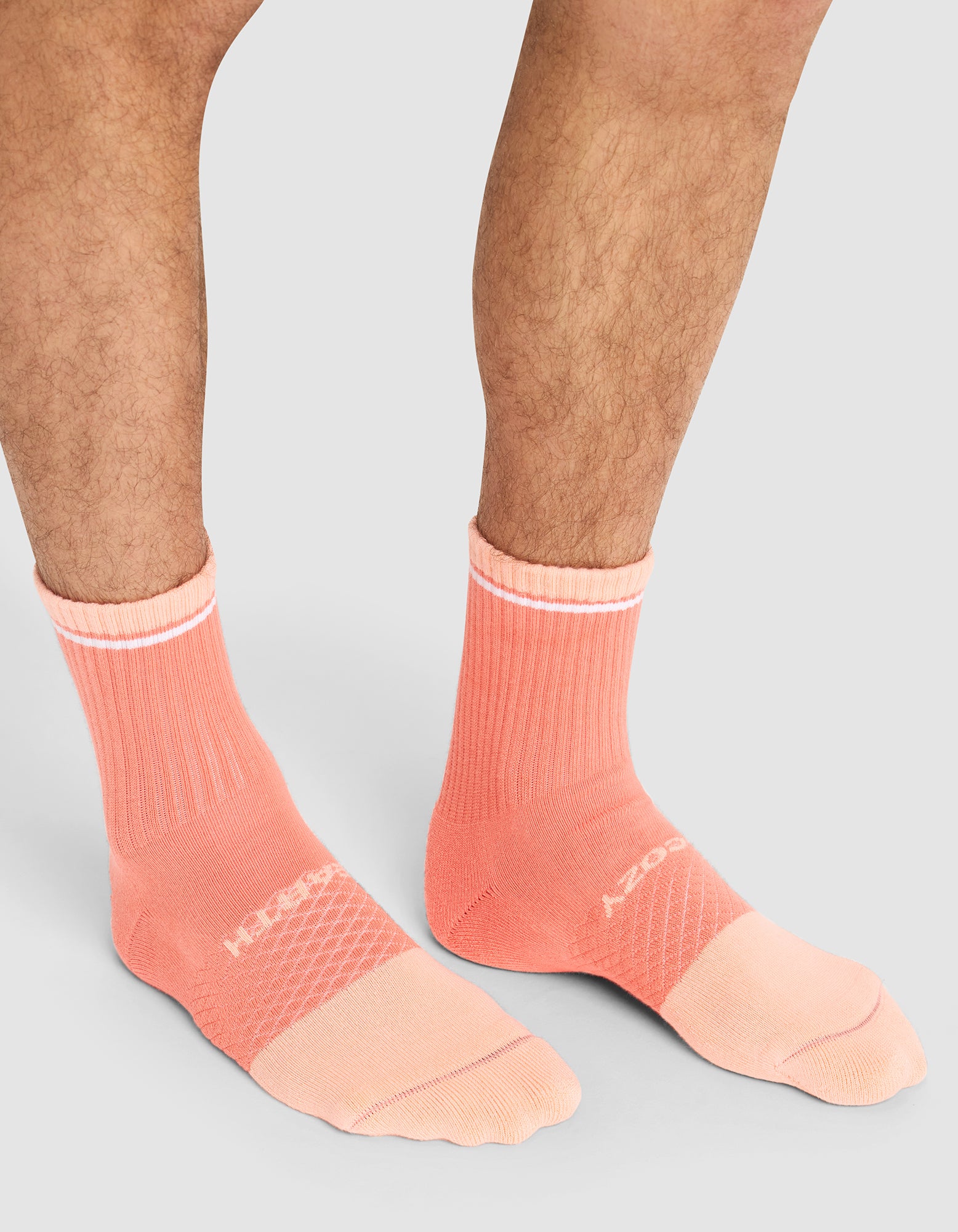 A person wears Cozy Earth Essential Calf Socks in peach with white text and accents, featuring ribbed cuffs and textured panels, standing on a light gray background. Only their lower legs and feet are visible.