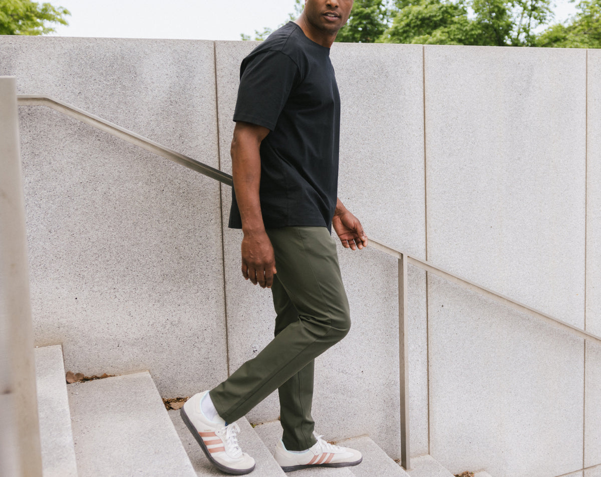 Man wearing a black t-shirt, green pants, and white sneakers standing against a concrete wall.
