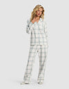 A woman with wavy blonde hair stands barefoot against a plain background, wearing the Cozy Earth Women's Farmhouse Long Sleeve Pajama Set—featuring a matching light-colored, plaid button-up top and loose-fitting pants. She gazes slightly to the side.|Color:Wintergreen Plaid