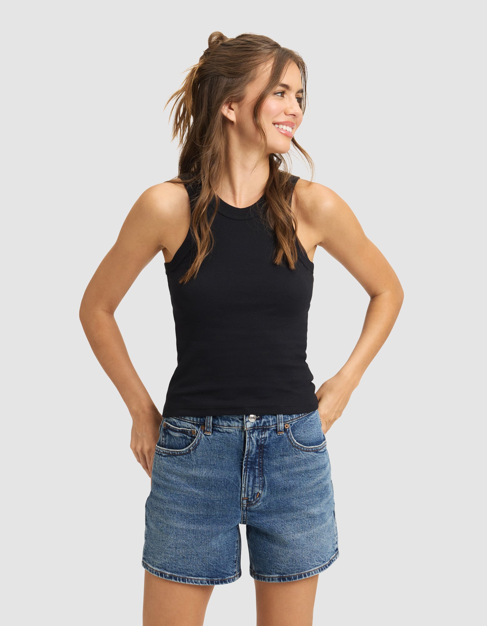 A woman with long brown hair smiles and looks aside, wearing the Cozy Earth Women's Fine Ribbed Tank in black and blue denim shorts. She stands with hands in pockets against a plain light background.|Color:Jet Black