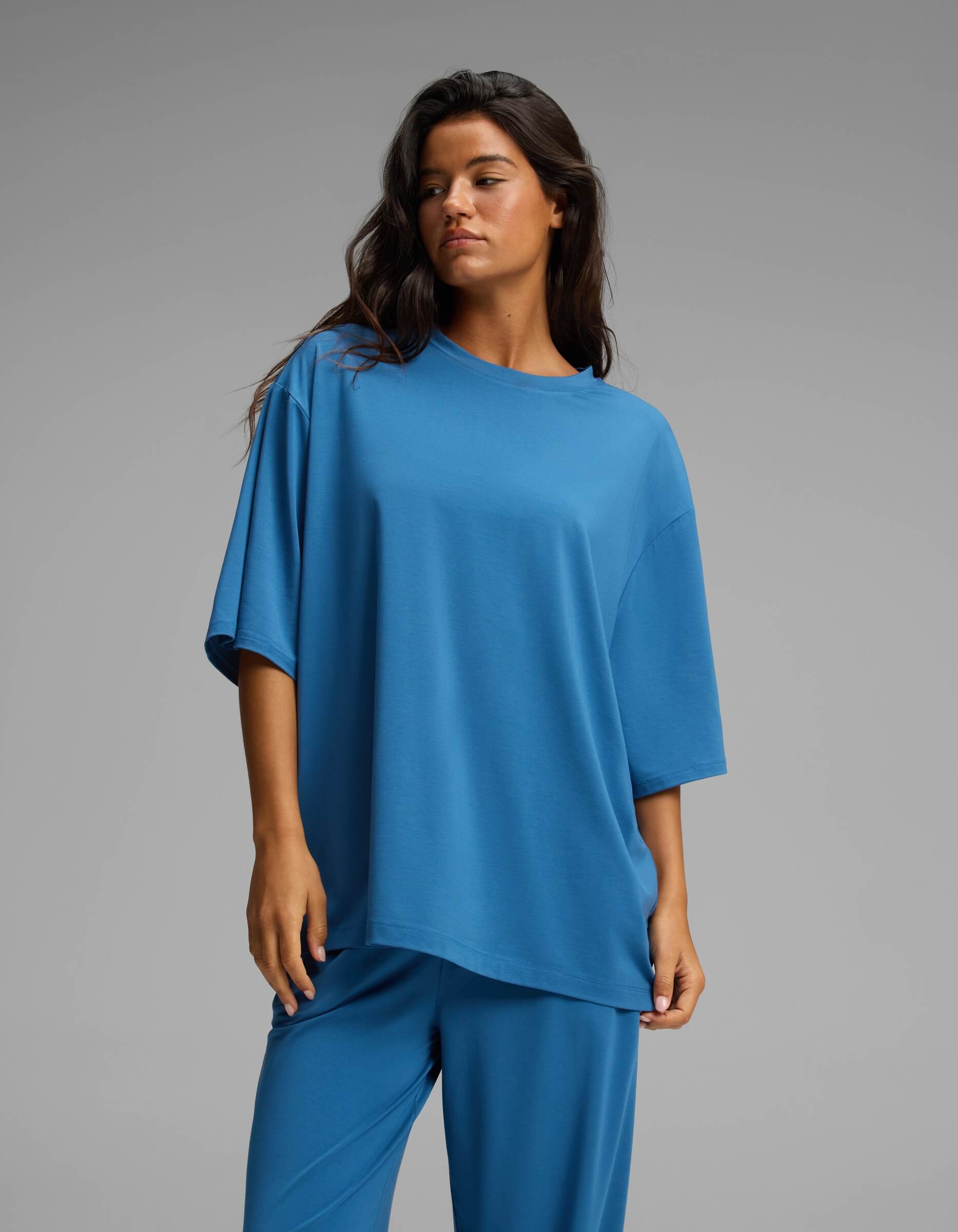A woman with long brown hair wears Cozy Earth’s Women’s First Layer Boyfriend Sleep Tee in blue, paired with matching pants. She stands against a plain gray background, gazing slightly to the side with a neutral expression.|Color:Anchor