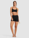 A woman stands barefoot against a plain background, wearing Cozy Earth's Women's First Layer Pajama Boxer Short in black and a matching black sports bra. She holds a rolled-up yoga mat in her right hand, looking forward with a relaxed expression.|Color:Jet Black
