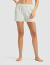 A woman stands barefoot in Cozy Earth's Women's First Layer Pajama Boxer Short and matching top, resting a hand on the waistband. The plain, light background highlights the light-colored, loose-fitting sleepwear set.|Color:Laurel