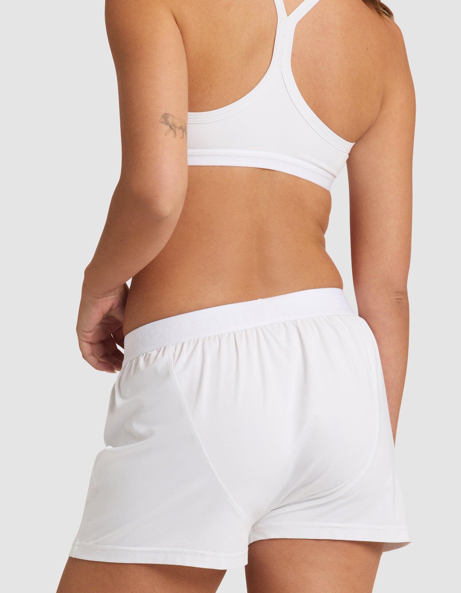 A woman is shown from the back against a plain light background, wearing Cozy Earth's Women's First Layer Pajama Boxer Short in white and a matching sports bra, with a small tattoo visible on her upper arm.