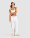 A woman with long brown hair in a ponytail stands casually against a plain background, arms crossed, wearing Cozy Earth's Women's First Layer Pajama Pant in white, paired with a white crop top and beige slippers.|Color:White