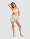 A woman poses barefoot against a plain background, wearing the Cozy Earth Women's First Layer PJ Soft Bra in a light shade with matching shorts. She has one arm bent, resting her hand on her head, and gazes at the camera.|Color:Laurel