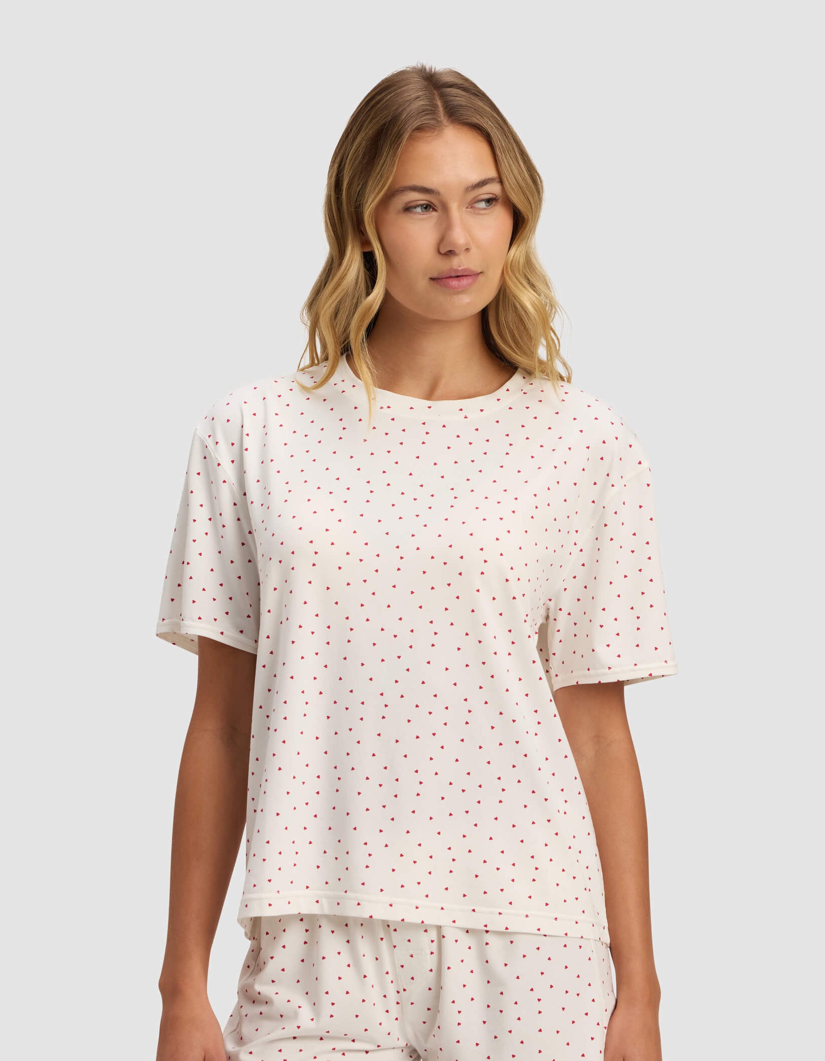 A woman wears the Cozy Earth Women's First Layer Pajama Short Sleeve Tee set with white fabric and small red polka dots, including a short-sleeve top and shorts, standing against a light gray background.|Color:Cranberry Sea Salt Mini Heart