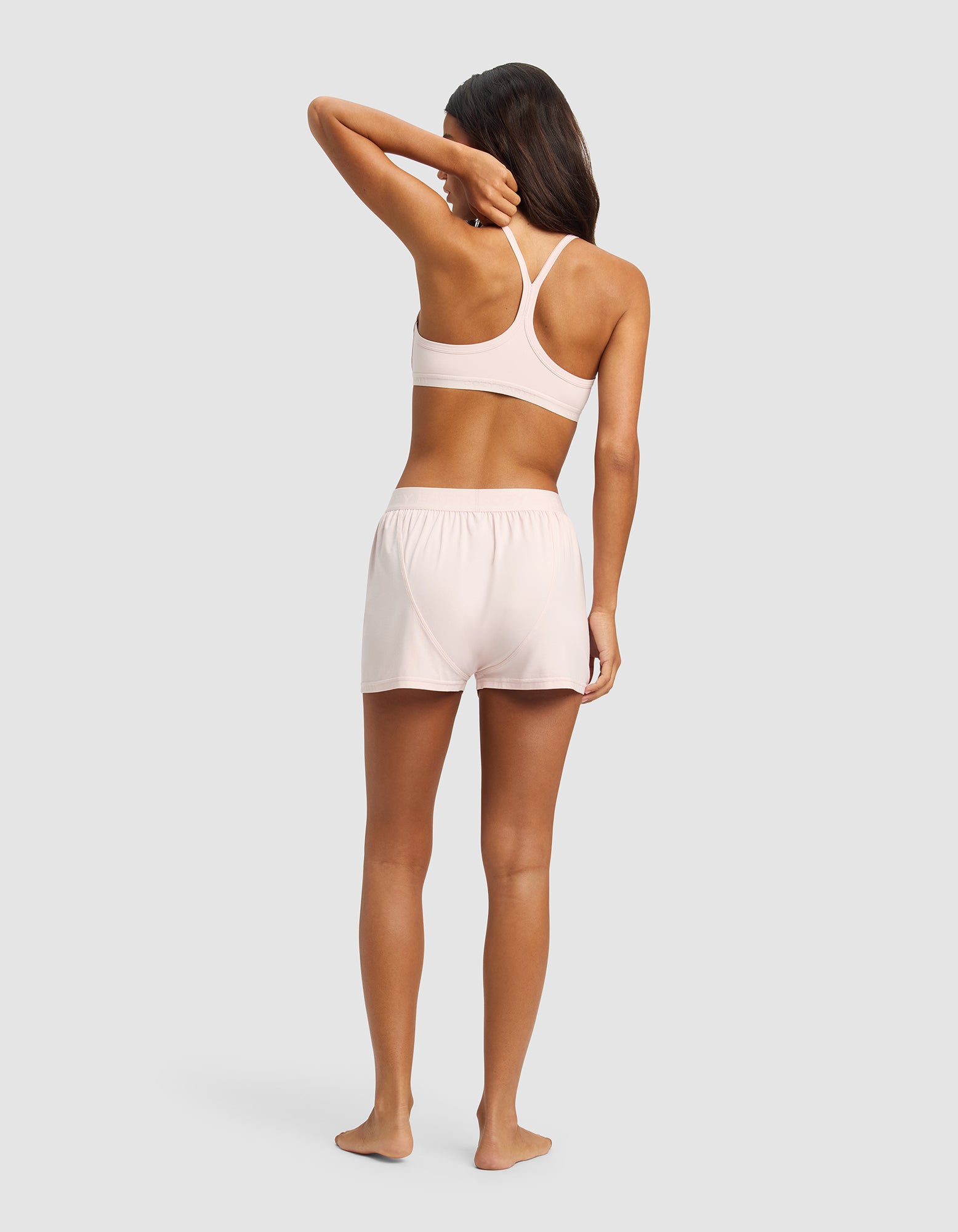 A woman stands barefoot on a plain background, facing away, wearing the Cozy Earth Women's First Layer Pajama Soft Bra & Boxer Short Set in light pink. One arm is bent with her hand touching her head, and her long brown hair is loose.