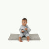 A baby sits barefoot on the Gathre Wholesale Padded Changing Mat in Stone Stripe, wearing a light gray outfit and holding a small object, set against a plain white background.