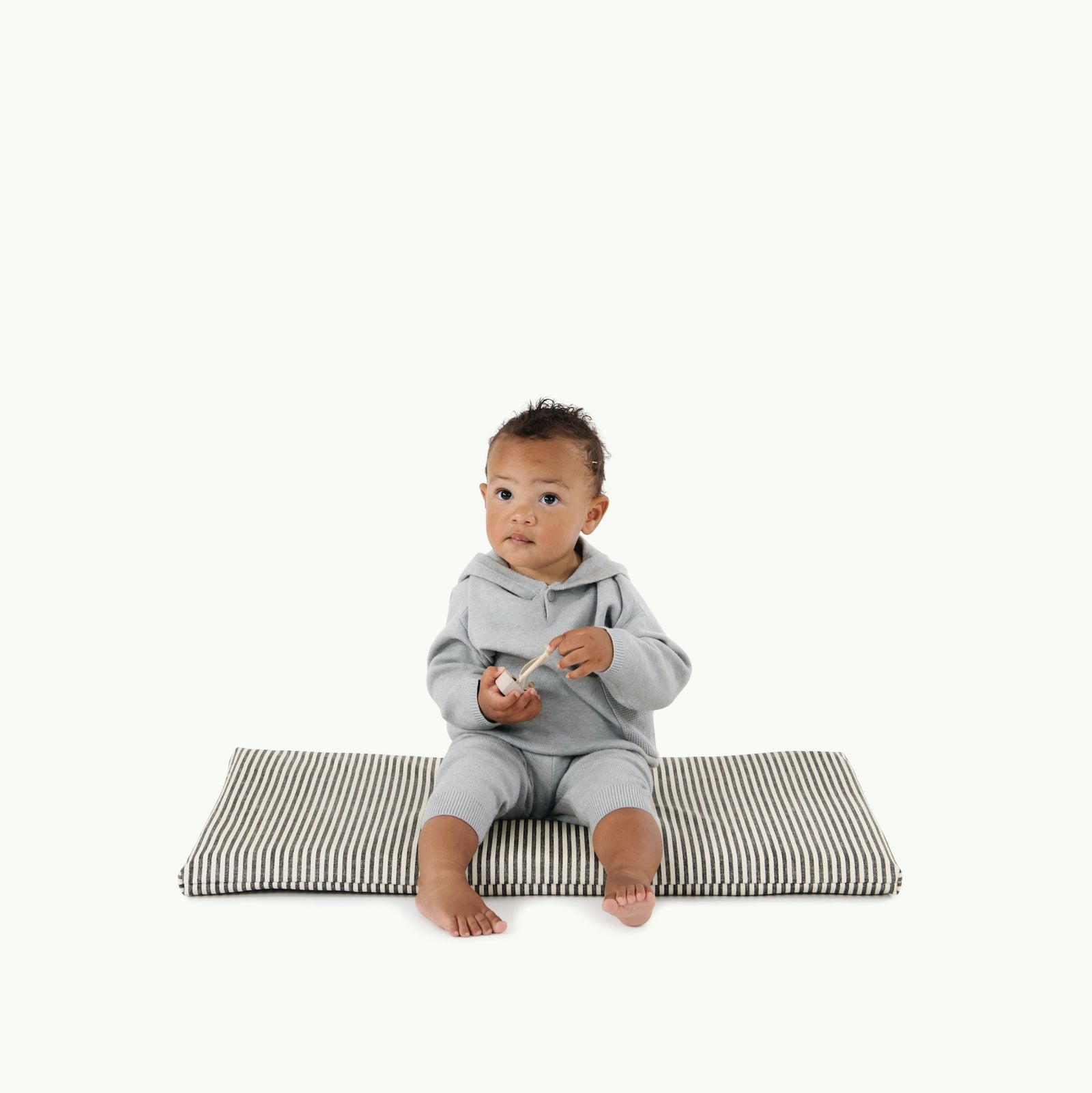 A baby sits barefoot on the Gathre Wholesale Padded Changing Mat in Stone Stripe, wearing a light gray outfit and holding a small object, set against a plain white background.